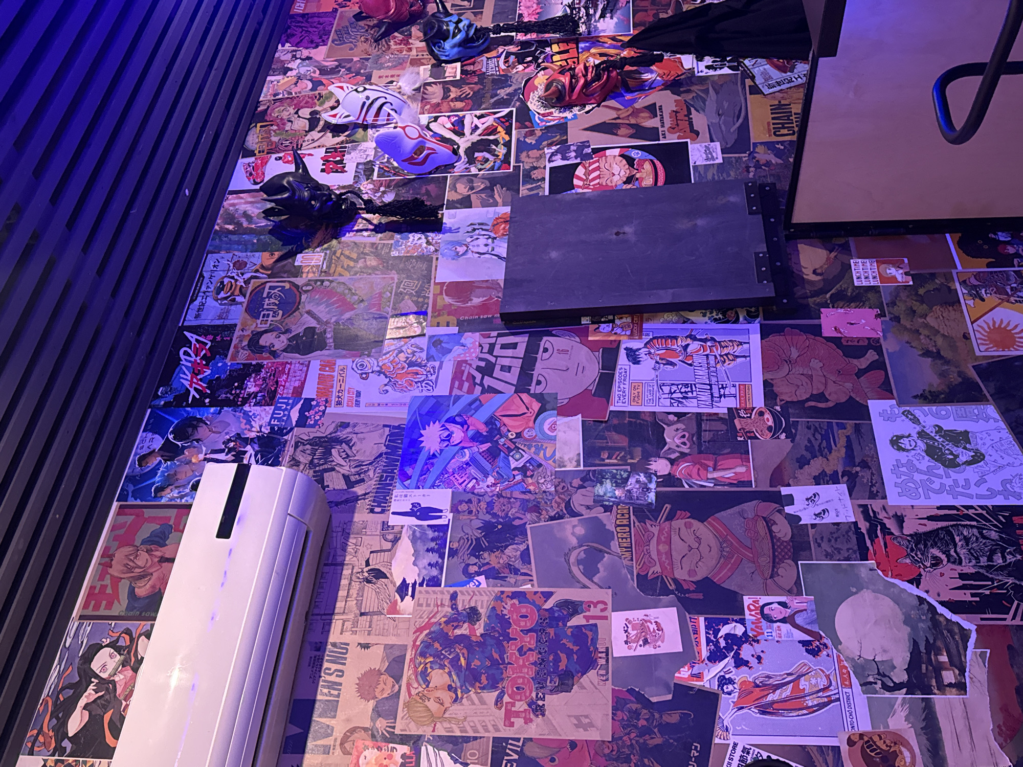 The image depicts a wall adorned with numerous anime-themed posters and masks. Various objects such as a black laptop-like device, a white air conditioner unit, and a table are positioned against the wall. The posters feature diverse anime characters and scenes, contributing to a vibrant and eclectic aesthetic. The lighting is notably bluish, casting shadows and enhancing the visual texture of the scene. The overall impression is that of an entertainment-themed setting, possibly a restaurant or bar, given the masks and table. There is no location found for "Unknown, Unknown."