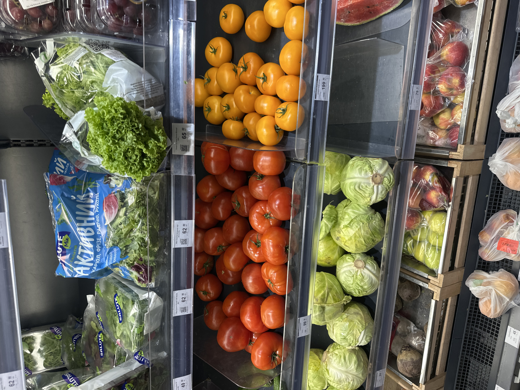 The image shows a supermarket aisle with a selection of fresh produce. In the foreground, on the bottom shelf, are bags of onions and what appear to be root vegetables like beets. Above this, on the middle shelf, are cabbages and a slice of watermelon. The shelf above that displays a variety of tomatoes, including red and yellow varieties. On the top shelf, there are packaged salads and lettuce. The overall environment is that of a well-stocked grocery store. There are no people visible in the image. The context of Ternopil, Ukraine is not visually evident from the image itself, as there are no specific landmarks or signage indicating this location.