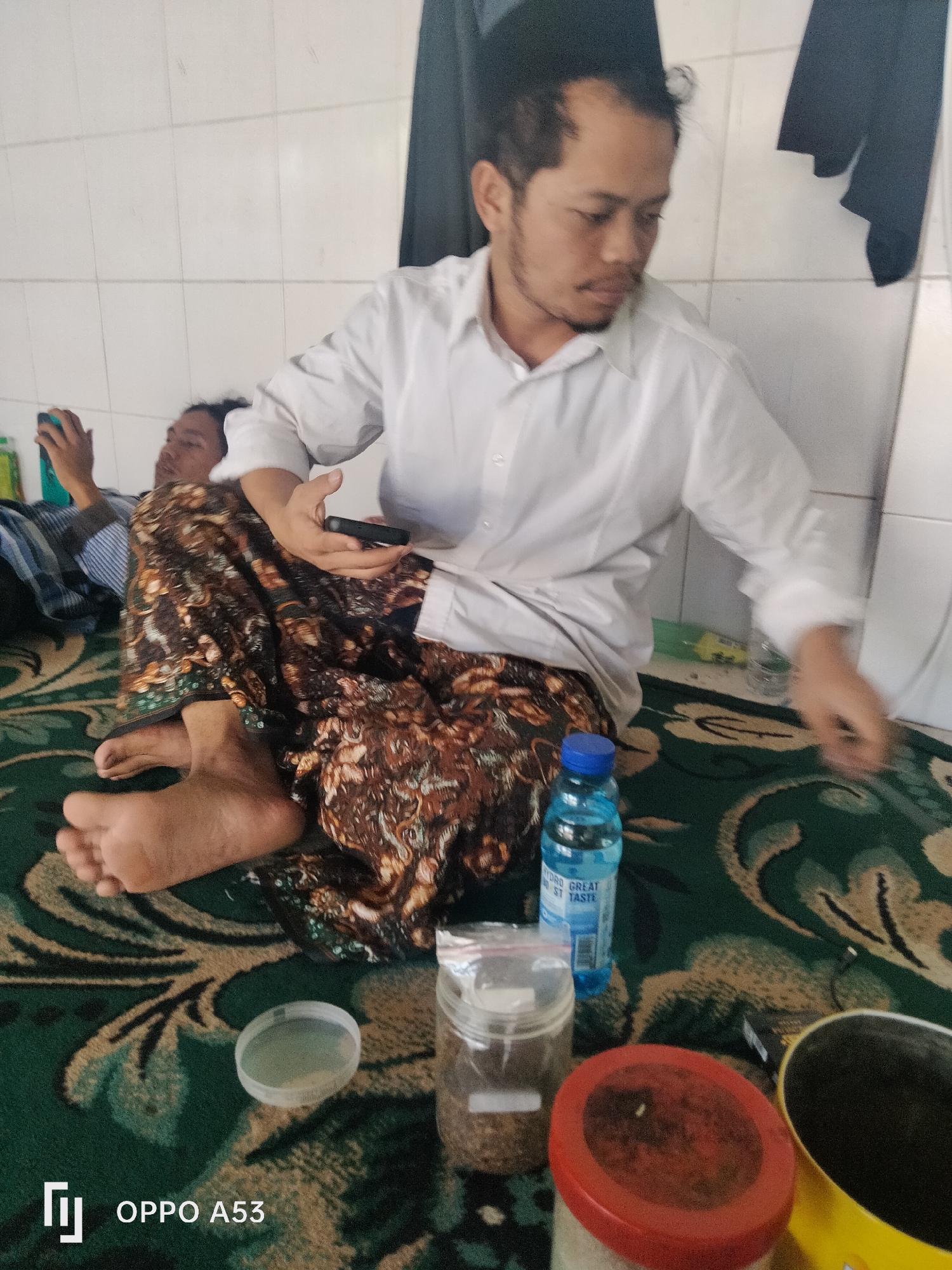 In a room with tiled walls, two men are seated on a green carpet with intricate patterns. One man, in the foreground, wears a white collared shirt and a patterned sarong. He is holding a black smartphone in his right hand. In front of him are a clear plastic bottle of "HYDRO GREAT TASTE" water, a jar with a white label containing a granular substance, a clear plastic container with a lid, and a yellow container with a black interior. The other man, in the background, is lying down and holding a blue object, possibly another phone. The image appears to have been taken in Badung, Indonesia.