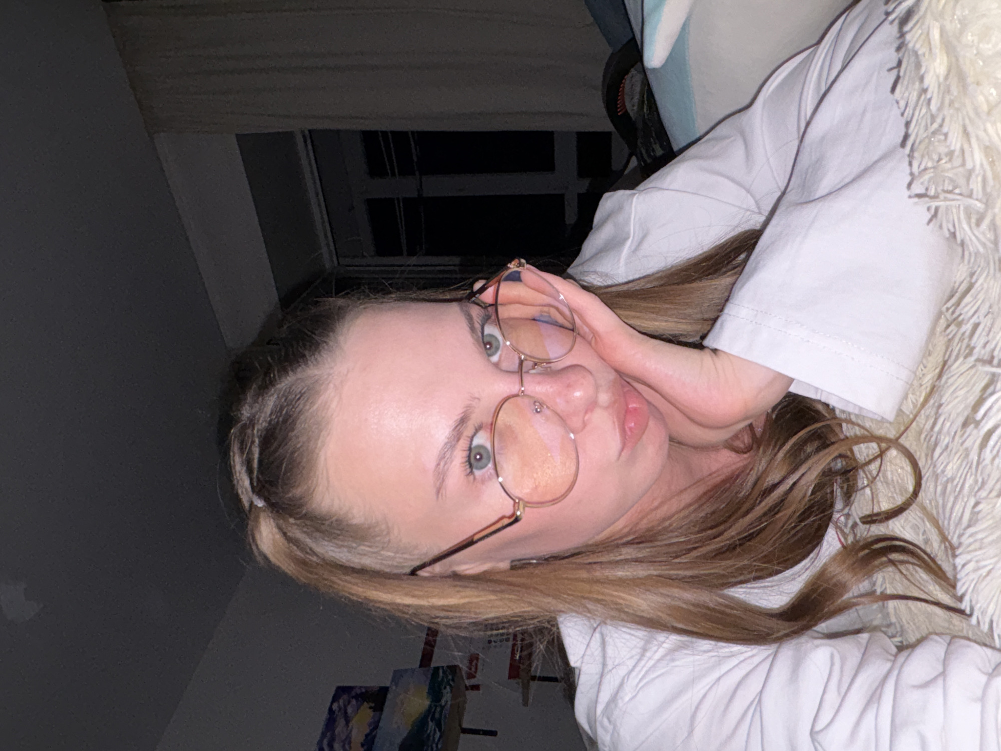 A young Caucasian woman with light brown hair styled in a ponytail, wearing glasses and a white shirt, is lying down and looking at the camera. She is resting her head on her hand, with her fingers touching her cheek. The background shows a room at night, with a window and curtains visible. There are also some paintings on the wall in the background. The scene appears to be indoors, possibly in a bedroom or living room. There is no information to suggest the location is the city of Perm, Russia.