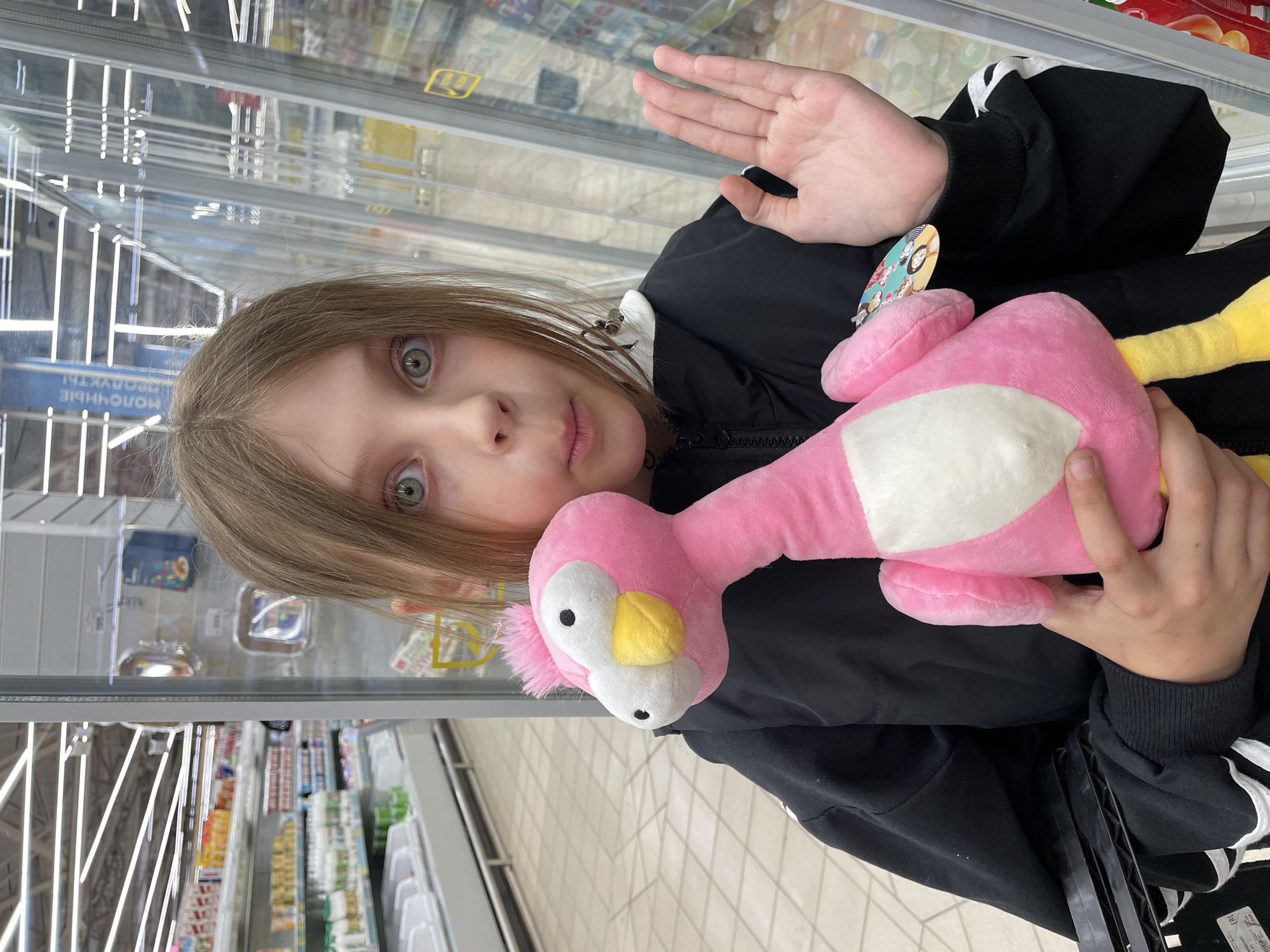 A young person with light brown hair and striking green eyes holds a pink flamingo plush toy with a white belly and yellow beak. The individual wears a black jacket with white stripes on the sleeves and stands in a supermarket aisle in Izhevsk, Russia. Glass-fronted refrigerator cases displaying various products are visible behind them. The person raises a hand, seemingly in a wave, while holding the flamingo in the other arm.