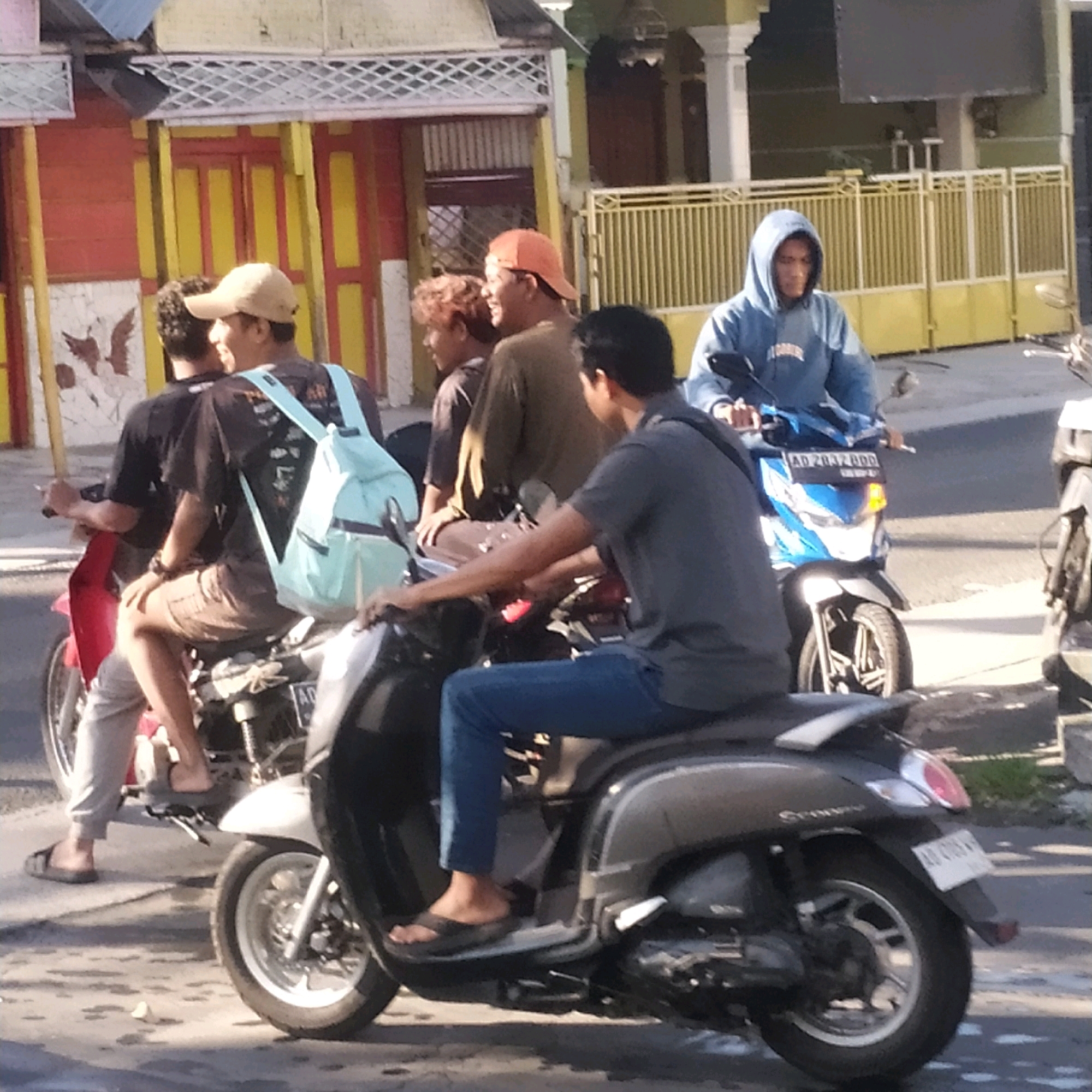 In Batan, Indonesia, on a sunny day, four individuals are observed outdoors. Two men are riding a red and black motorcycle. The man in front is wearing a brown shirt and a tan baseball cap, while the man behind him has a black shirt and a light blue backpack. They are looking in different directions. A third man, wearing a blue hooded sweatshirt, is on a blue and white motorcycle to the right, looking ahead. A fourth person, partially visible, is seated on a black scooter in the foreground, wearing a grey shirt and blue jeans, with their hands on the handlebars. The background shows a building with yellow and red accents, a yellow fence, and some greenery. The ground is paved with some wet patches.