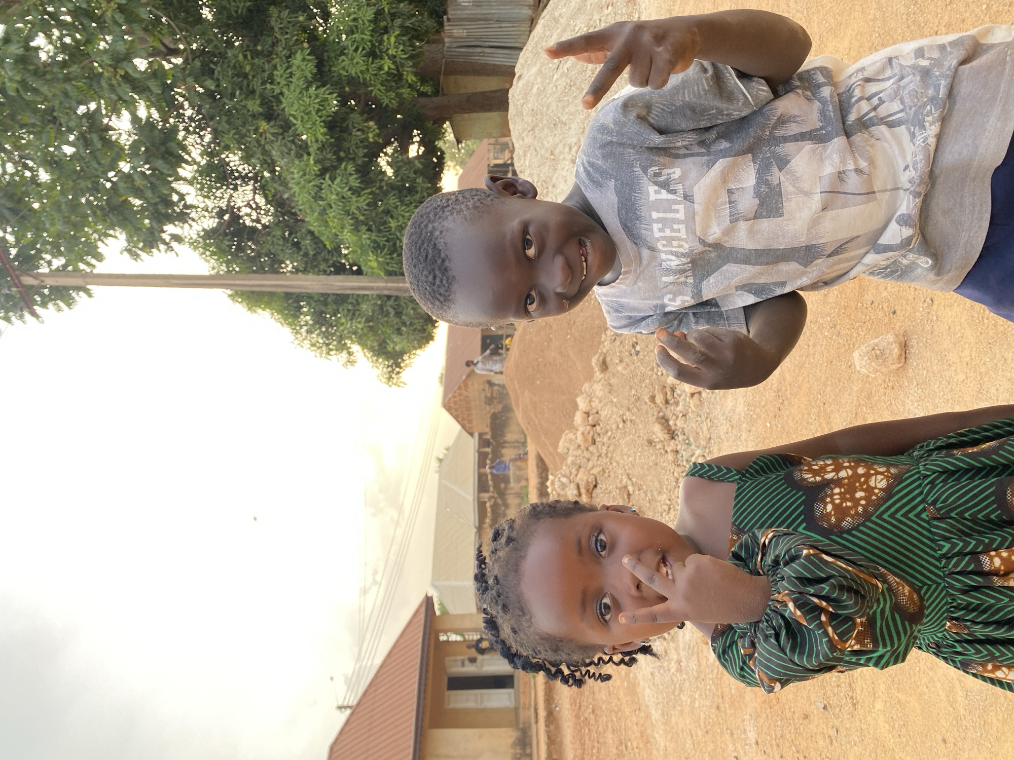 Two children, a boy and a girl, are photographed outdoors in Kuru Dungus, Nigeria. The boy wears a patterned t-shirt and the girl wears a green and brown patterned dress. Both children are making a hand gesture. In the background, there is a building and a pile of dirt. Trees are also visible in the background. The ground is dirt or sand.