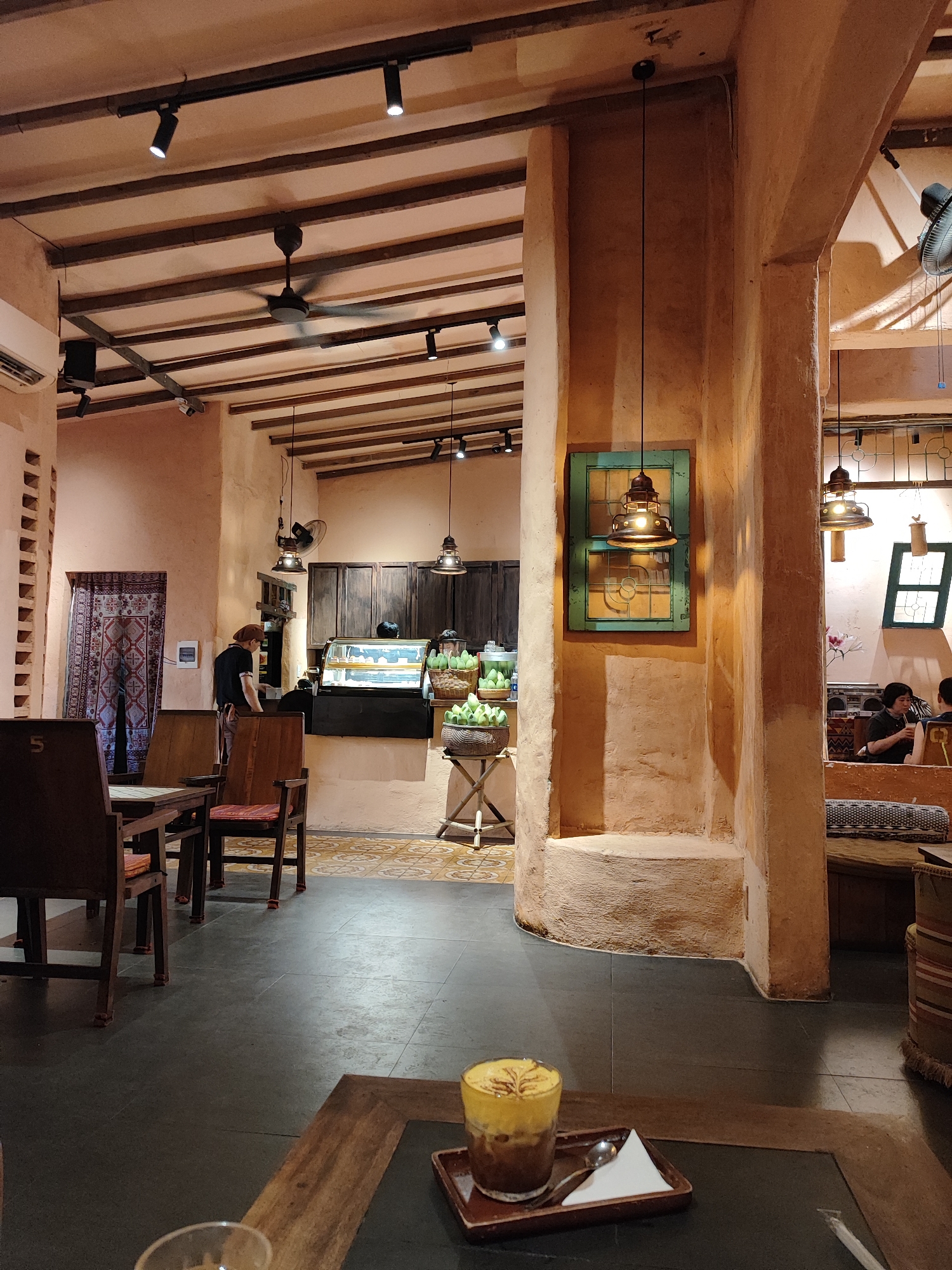 This image depicts the interior of a cafe in Da Nang, Vietnam. The cafe has a rustic and traditional aesthetic, with exposed wooden beams on the ceiling and rough, textured walls. A ceiling fan is visible, along with track lighting and decorative pendant lamps.
In the foreground, a glass of iced coffee with foam art sits on a wooden table. To the left, wooden chairs and tables are arranged.
Further into the cafe, a counter area is visible with a display case showing pastries or food items. A person wearing an apron and a head covering is behind the counter. To the right of the counter, there are baskets of green fruits, likely mangoes.
In the background, there are seating areas with upholstered benches and cushions, and several people are seated and interacting. Decorative elements like a tapestry on the wall and framed window panes add to the ambiance. The floor is tiled with a pattern. The overall impression is of a cozy and authentic Vietnamese cafe.