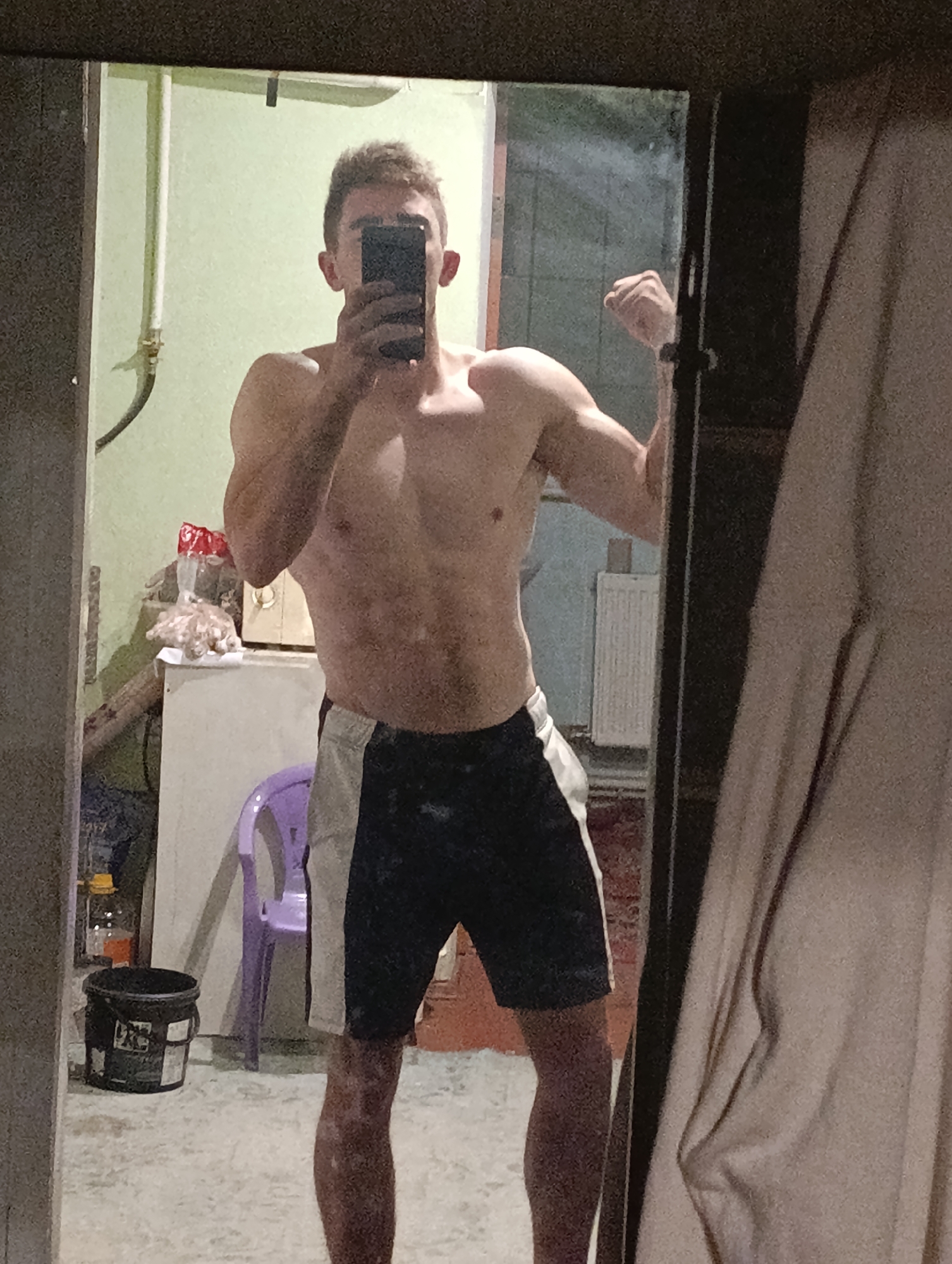 The image shows a shirtless young man in gym shorts standing in front of a mirror, taking a selfie. He is flexing his bicep. In the background, there is a purple chair, a black bucket, and some shelves with items on them. The overall environment appears to be indoor, possibly a room in a house or a gym. The image does not provide any specific information or visual cues that would place the scene in Fastiv, Ukraine.