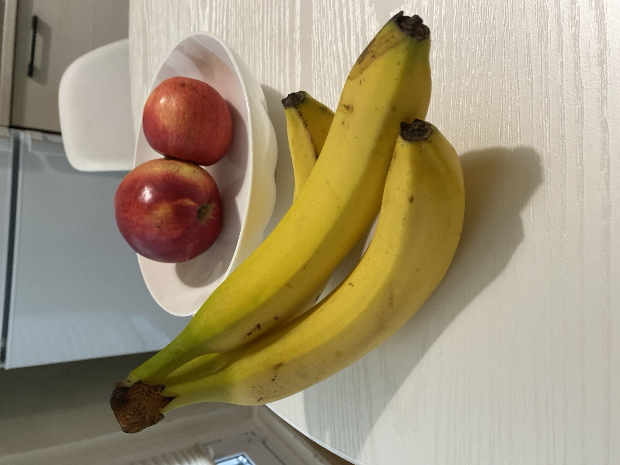 A bunch of three ripe bananas lies diagonally across a white wooden table. To the upper right of the bananas, two red and yellow apples sit in a white bowl. In the background, a white refrigerator and a white chair are visible. The scene is indoors, likely in a kitchen.