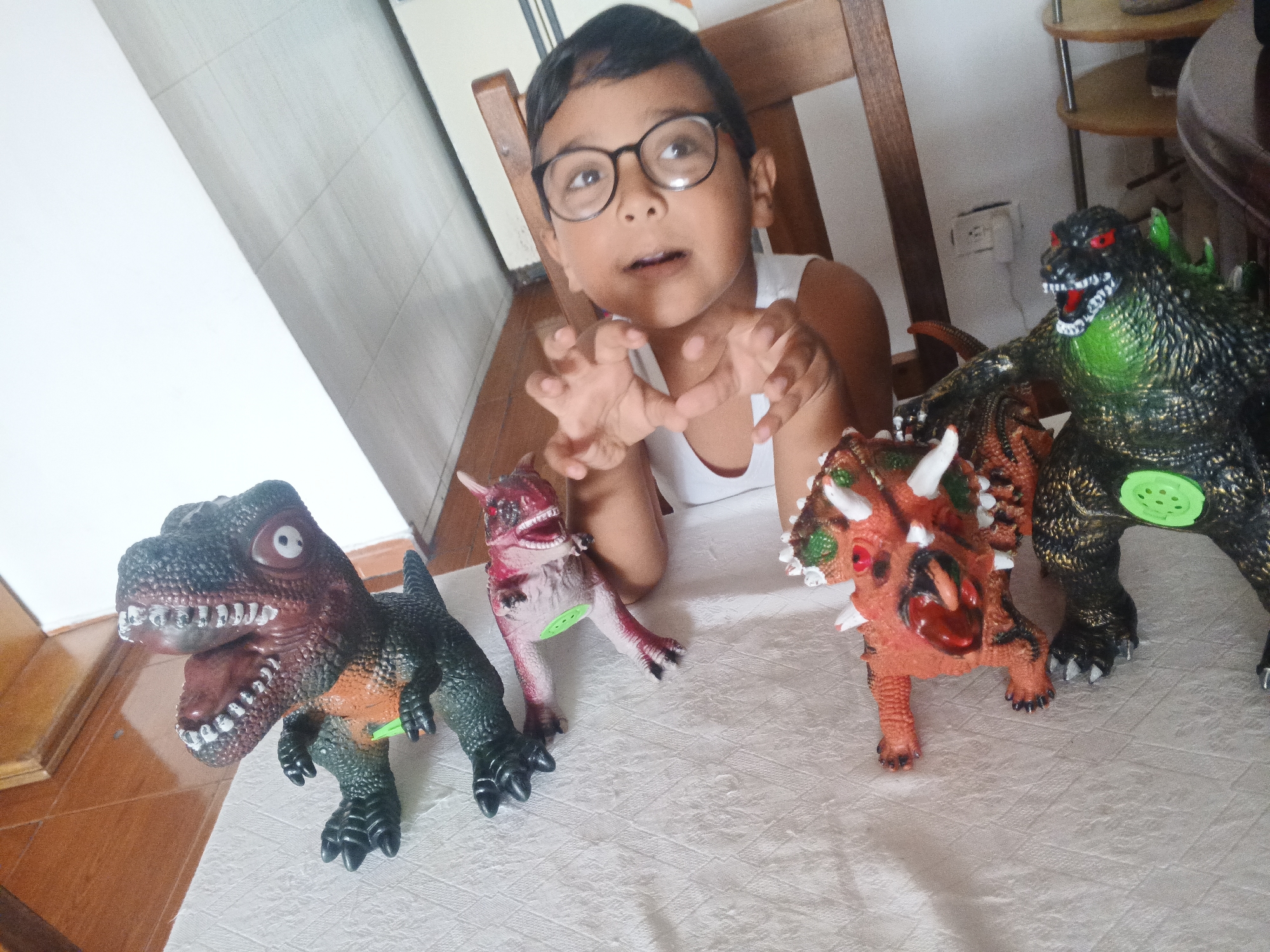 A young boy with dark hair and glasses, wearing a white tank top, is captured in an indoor setting, deeply engaged in playing with four dinosaur toys. He is leaning forward with his hands raised in a claw-like gesture, and his mouth slightly open, suggesting an imitation of the roaring dinosaurs or an expression of excitement.
The four distinct dinosaur toys are prominently displayed on a white, textured surface, possibly a tablecloth. From left to right, they include: a dark green and orange Tyrannosaurus Rex-like figure with an open mouth revealing teeth; a smaller pink and red dinosaur with a wide-open mouth; an orange and brown Triceratops-like toy featuring white horns and red eyes; and a large, dark green and black Godzilla-like figure with red eyes and sharp teeth, which also has green spikes on its back. Some of the toys have visible green circular or protruding elements on their sides or bellies.
In the background, parts of a wooden chair and a wooden table or shelf are visible. A white electrical wall outlet is present on the light-colored wall behind the toys on the right side of the frame. The lighting suggests it is daytime. The overall scene depicts a moment of joyful and imaginative play. There is no visible text in the image. The location is contextualized as Caracas, Venezuela.