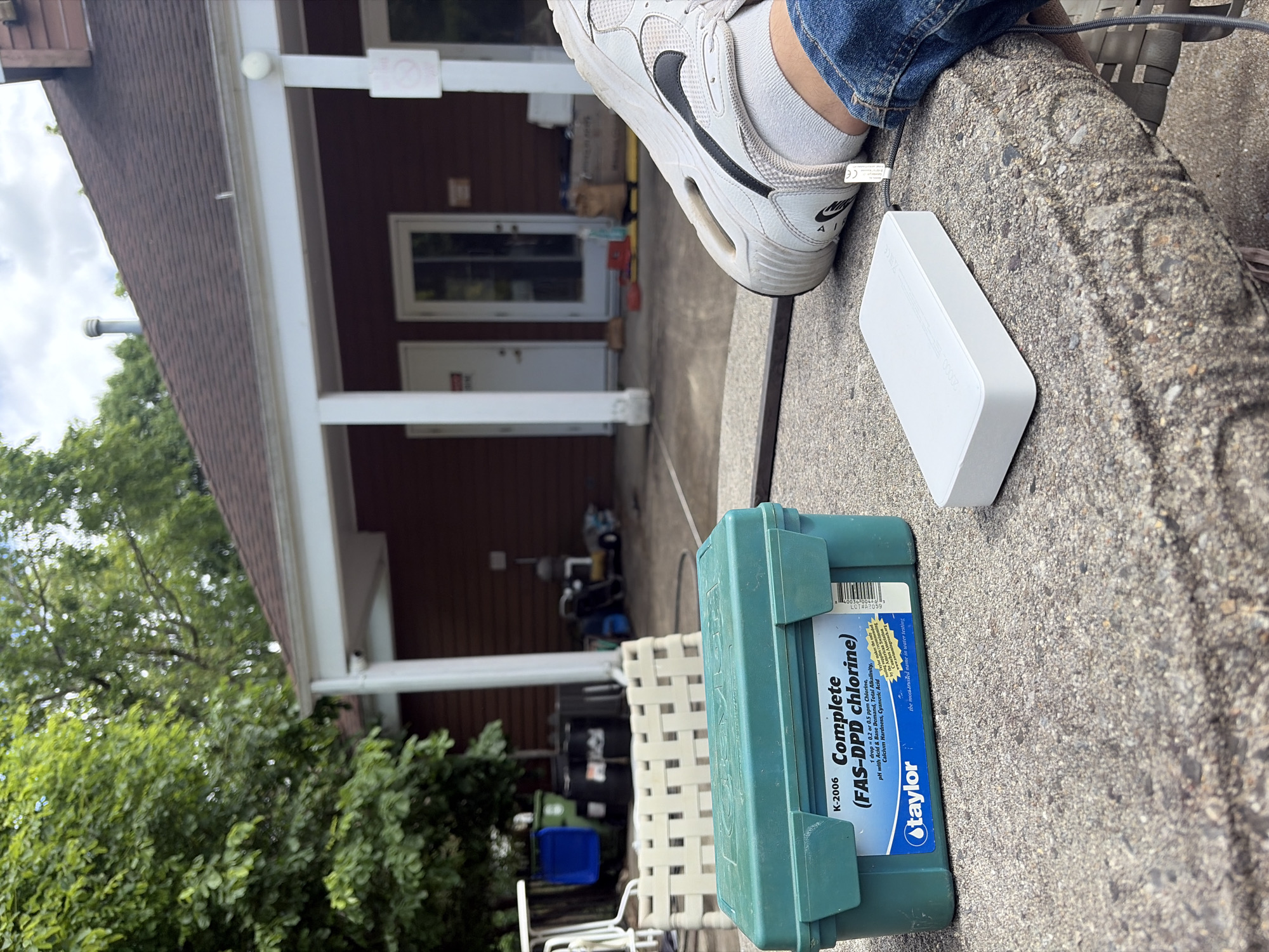 The image shows an outdoor setting, likely a backyard or patio area. A leg with a white sneaker and blue jeans is visible in the upper right corner. A white rectangular object, possibly a power bank or similar electronic device, is positioned next to the sneaker. Below the white object is a green container labeled "Taylor Complete (FAS-DPD chlorine) K-2006". The label indicates it's a pool water testing kit. In the background, a house with a brown exterior and white trim is visible. A white plastic chair is partially visible near the testing kit. Green trees are seen on the left side of the frame. The photo could possibly be located in Edison, New Jersey.