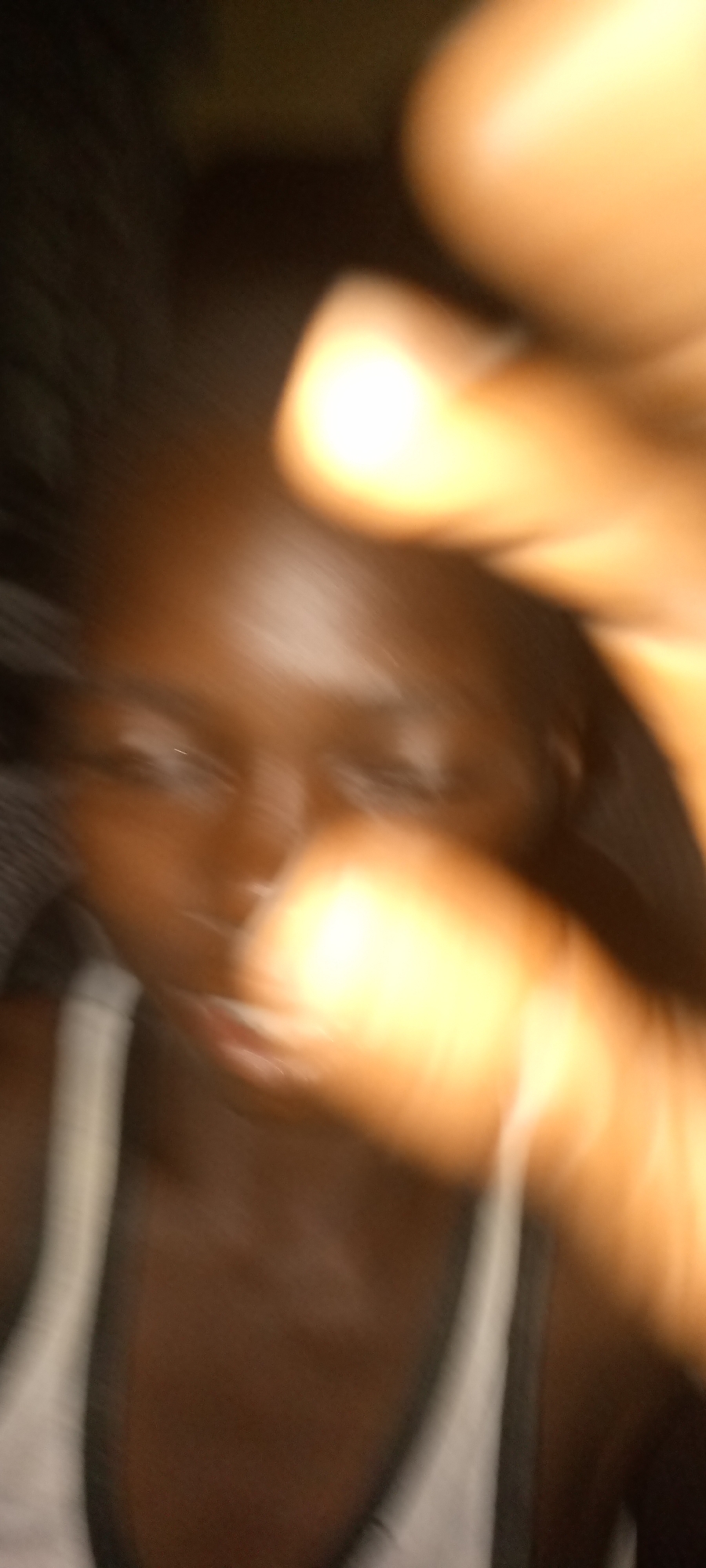This is a highly blurred and close-up image, giving the impression of a spontaneous selfie or a candid shot taken with movement. The central subject is a person, likely a young individual, with dark skin and dark hair that is partially visible on the left side. Their face is largely obscured by extreme motion blur and bright, warm-toned light, which appears to emanate from a hand or fingers held very close to the camera lens in the foreground, casting an intense golden-orange glow across the right and central portions of the image.
The person's eyes are faintly discernible, but their expression is indistinct due to the blur. They appear to be wearing a light-colored (possibly white) tank top or camisole, with a darker strap visible on their left shoulder. The background is very dark and indistinct, offering no specific location cues, though it suggests an indoor or dimly lit setting. No specific activity beyond posing for a photo can be identified. The lighting and blur make it difficult to determine the exact time of day, but the presence of bright, warm light sources implies artificial lighting. There is no visible text in the image.