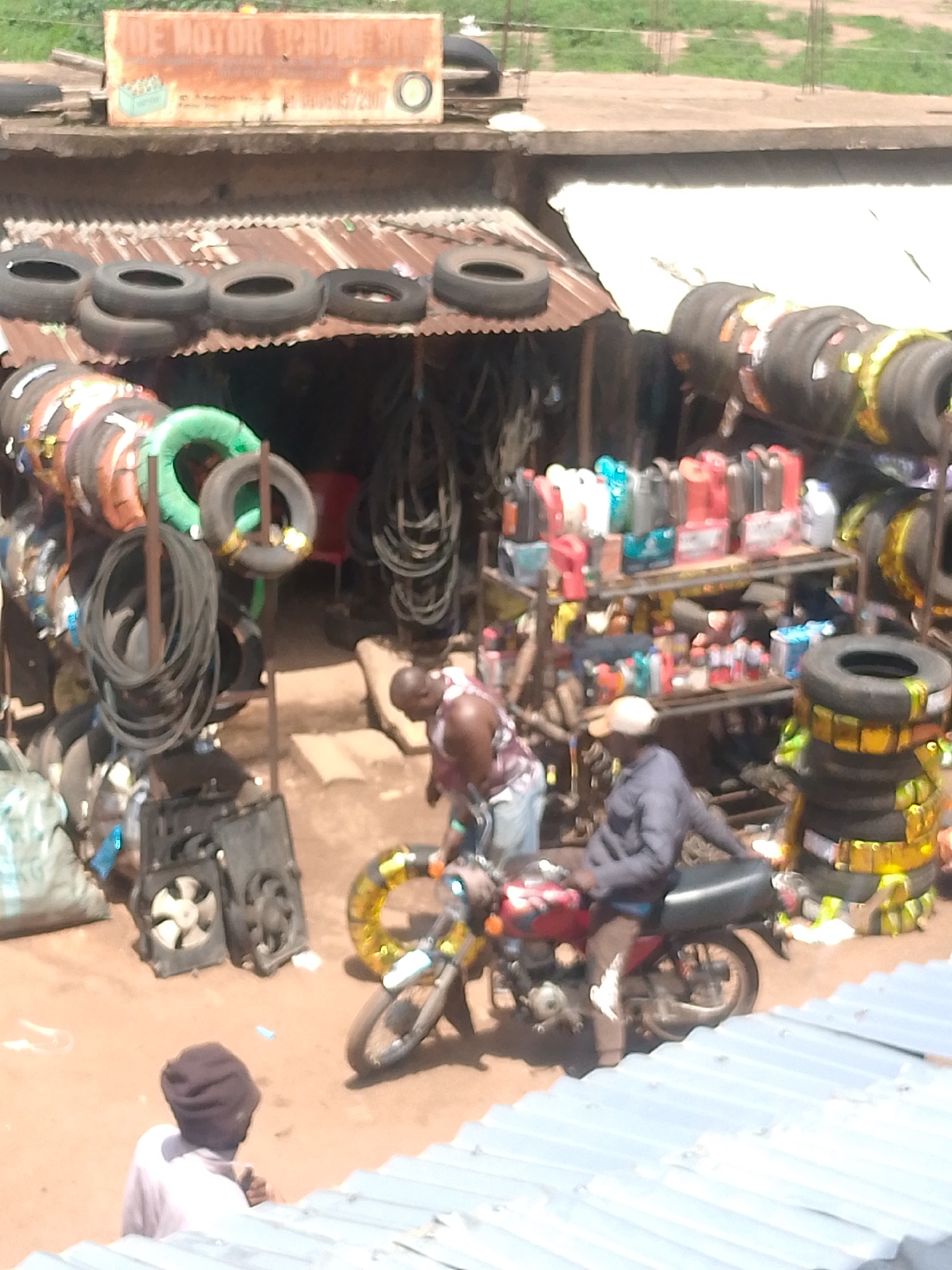 The image depicts a roadside tire shop in an urban environment, possibly in a city named Unknown, Unknown. The shop is open-air with a corrugated metal roof, where several tires are stacked. More tires are displayed around the shop, along with other automotive parts, including liquids and what appears to be car radiators. Two men are seen working on a red motorcycle; one is crouched down near the front wheel, while the other is seated on the bike. A third man is standing nearby, facing away from the camera. The ground is dusty and appears to be unpaved. The overall setting suggests a low-income or developing area.