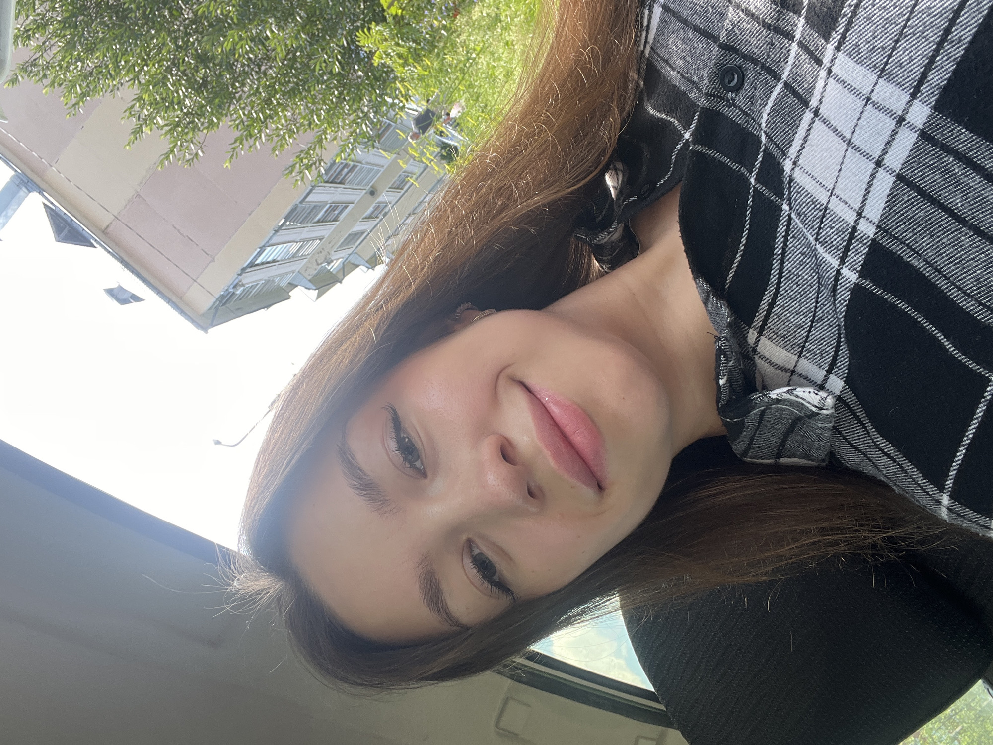 A young woman with long brown hair, wearing a black and white plaid shirt, takes a selfie outdoors. Her face is turned towards the camera, and she is smiling. She has on light makeup and appears to be in a car, as indicated by a portion of a black car seat visible on the left side of the frame. In the background, a light-colored building with multiple windows and a green tree are visible against a bright, overcast sky. The overall scene suggests a casual outdoor setting within a city.