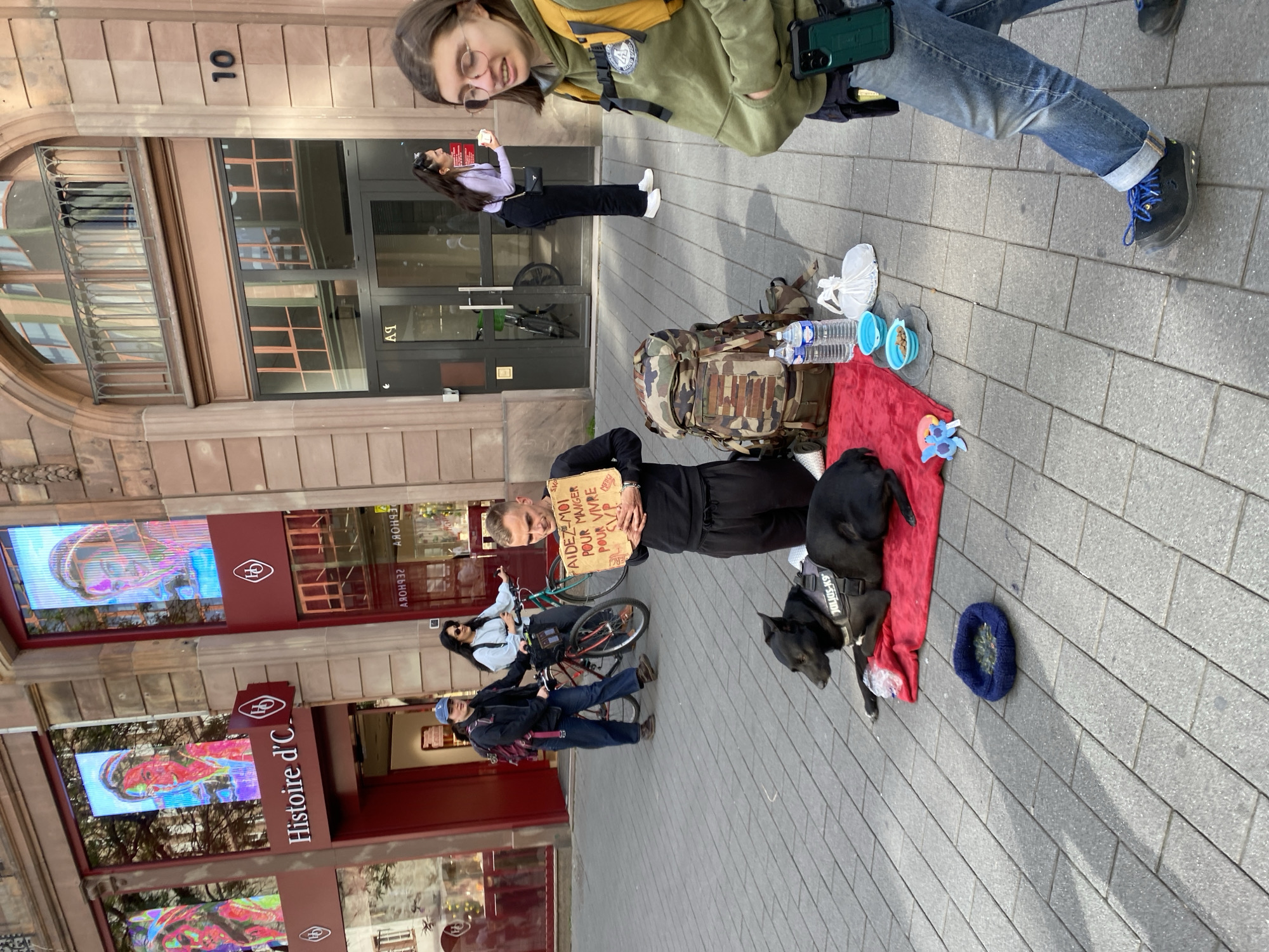 The image depicts a street scene in Strasbourg, France, centered on a man and his dog, suggesting a situation of begging or homelessness. The man, with short hair, stands holding a hand-drawn cardboard sign that reads "AIDEZ-MOI POUR MANGER POUR VIVRE S.V.P." (Help me to eat, to live, please). He is dressed in dark clothing.
Beside him, a black dog, possibly a Labrador mix, lies calmly on a red blanket. The dog wears a harness with "JULIUS-K9" visible on it. Arranged on the grey paved sidewalk next to the dog are a camouflage backpack, two bottles of water, two blue collapsible dog bowls, and a small blue dog bed.
In the background, several pedestrians are visible. A young woman in a green hoodie and glasses is in the immediate foreground, looking down. Another woman in a light purple top and black pants stands further back near a building entrance marked with a reversed "A9". A person on a bicycle and another individual in a blue cap are also seen in the mid-ground.
The setting is an urban street lined with multi-story buildings. Shop windows feature large digital displays showing colorful, abstract portraits, and one shop is clearly marked with the sign "Histoire d'C". Building numbers, such as "10", are visible on the facade, and reflections in windows show reversed text including "SEPHORA". Bright daylight is indicated by the clear shadows cast on the pavement, suggesting clear weather. The overall scene portrays a moment of daily life on a bustling city street.