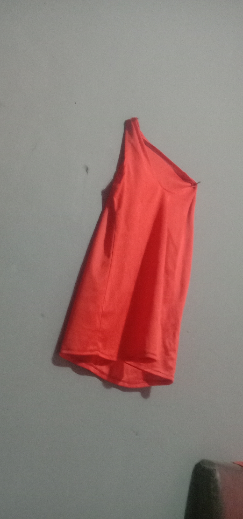 An orange, one-shoulder top hangs on a grey wall. The top is made of a soft material and has a flowy silhouette. It is slightly wrinkled, suggesting it may have been recently worn or is in storage. The wall is bare except for a few faint marks and a shadow cast by the garment. In the lower right corner of the image, a portion of a dark brown chair is visible. The lighting in the scene appears to be artificial, creating a somber mood. The location is unspecified within Kendari, Indonesia, and there are no people present.