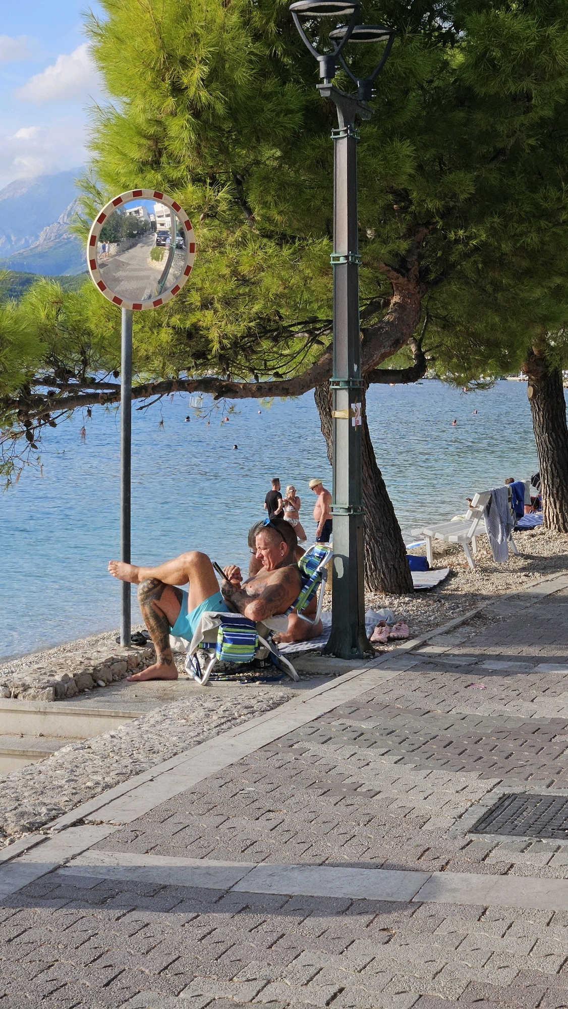 This image depicts a sun-drenched scene at a beach in Općina Tučepi, Croatia. In the foreground, a man with extensive tattoos is seated in a striped beach chair, looking at his phone. He is wearing blue swim trunks. His bare feet are visible, and one leg is crossed over the other. Next to him, another person, also tattooed, is reclining on a mat. The beach is made of small pebbles and sand, transitioning to a paved walkway. A lamppost stands nearby, casting a shadow.
In the background, the clear turquoise water of the Adriatic Sea is visible, with several people swimming and wading. Further inland, lush green pine trees provide shade, with their branches extending over the beach area. A white bench with towels draped over it is situated on the pebble beach. A convex traffic mirror, mounted on a pole, reflects a road with cars and a few pedestrians. The sky is blue with scattered white clouds, and a mountainous landscape is visible in the far distance. The overall atmosphere is one of leisure and relaxation.