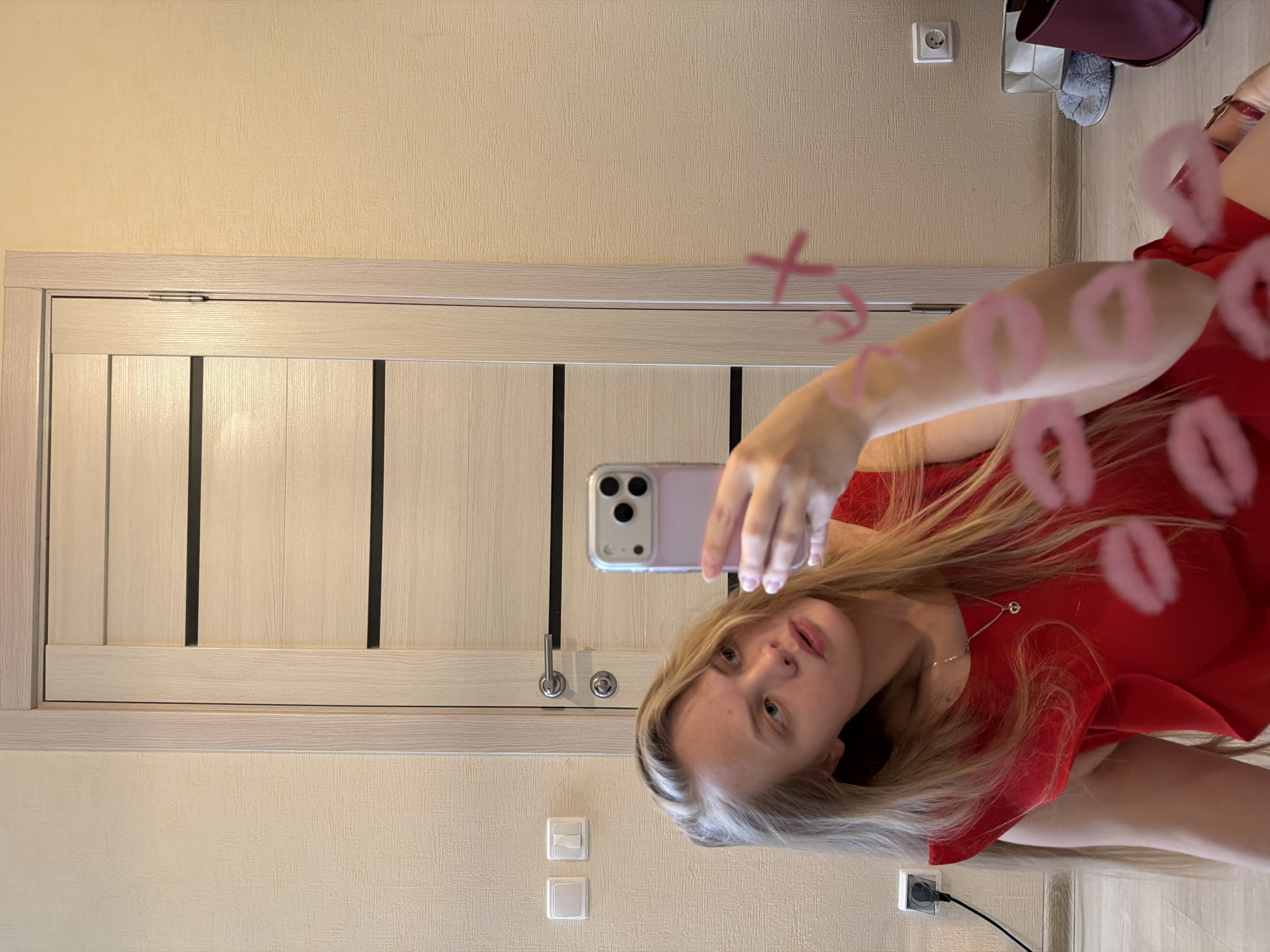 A young woman with long, light blonde hair is taking a selfie in front of a mirror. She is wearing a red dress and a silver necklace. The mirror has pink kiss marks and an "XO" written on it. In the background is a wall painted in a light beige color, a white door with a grey frame, and an electrical outlet. There is also what appears to be a purse and shoes. Based on the context, this photo was likely taken in a private residence in Izhevsk, Russia.