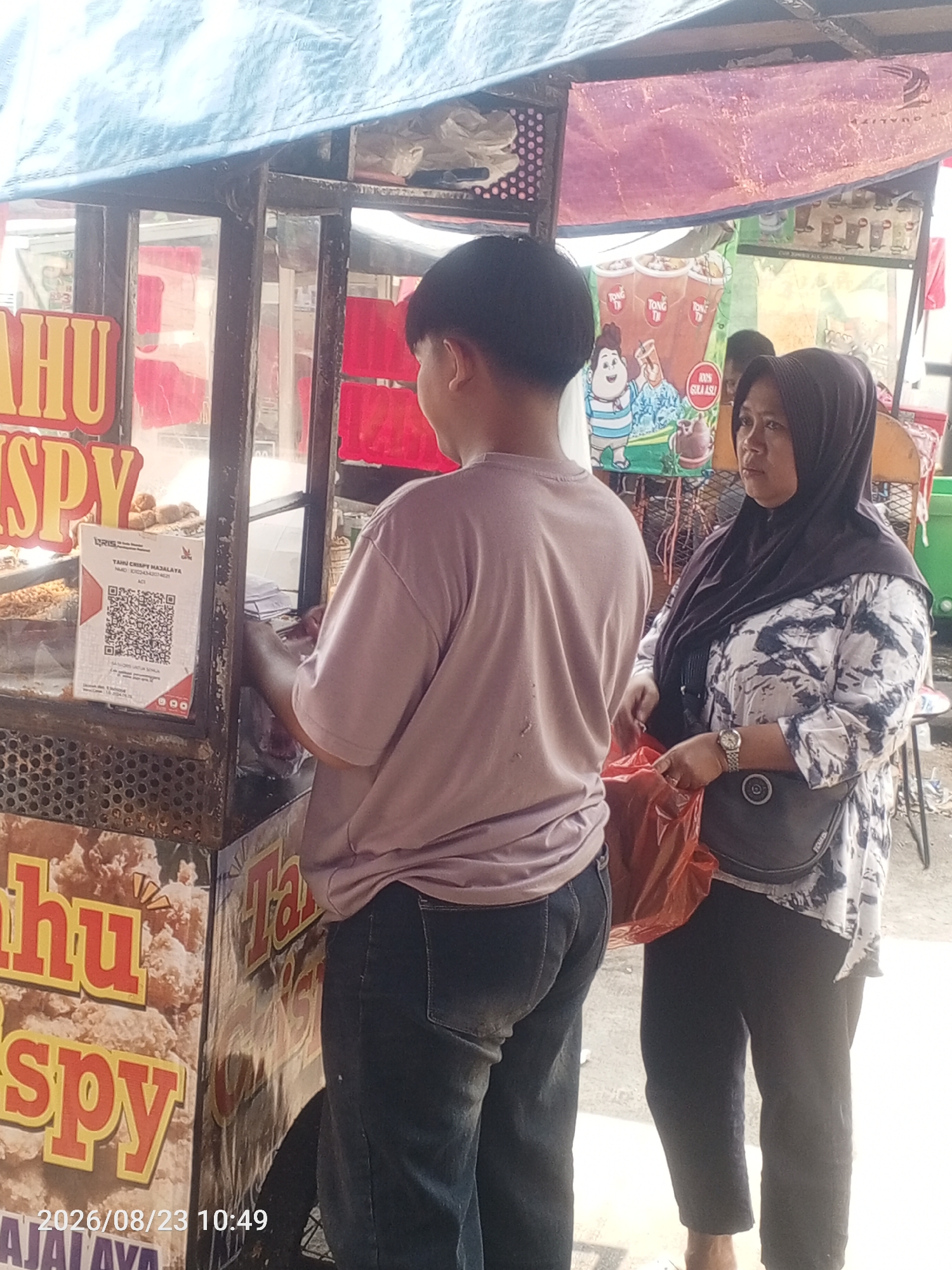 Two people are standing in front of a food cart in Depok, Indonesia. A young person wearing a light purple t-shirt and dark jeans stands on the left, facing away from the camera and looking at the food cart. On the right, a woman wearing a black hijab and a patterned black and white blouse stands, holding a bright orange plastic bag and looking towards the food cart. The food cart has signs with text in Indonesian, including "HU CRISPY" and "TAHU CRISPY MAJALAYA". A QR code is displayed on the cart. The background shows a partially obscured awning and other elements of a street market or food stall. The timestamp "2026/08/23 10:49" is visible in the bottom left corner.