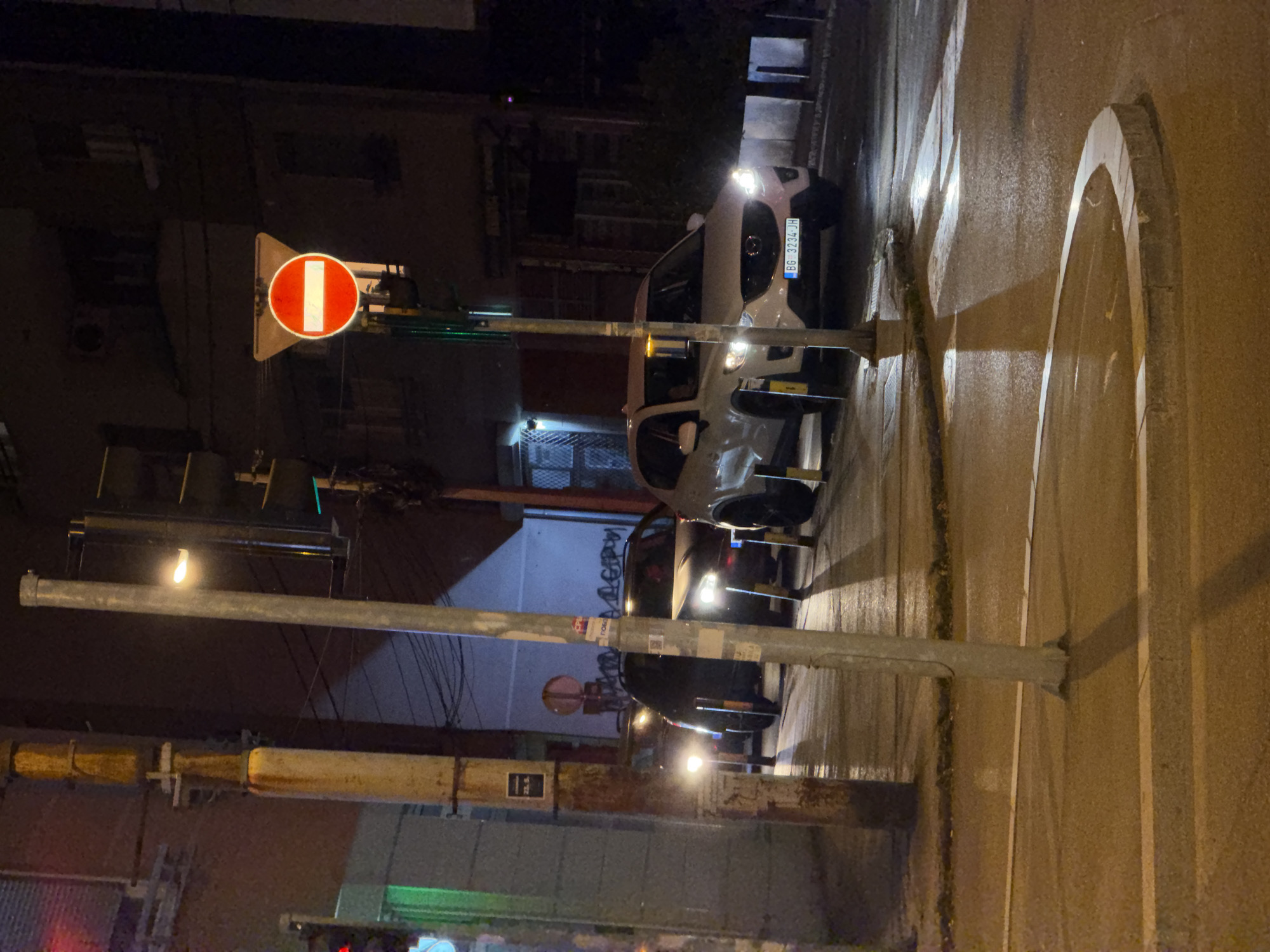 The image depicts a street scene at night in Vracar Urban Municipality, Serbia. A "Do Not Enter" traffic sign is visible on the left side of the frame. A parked white car has a license plate that reads "BG 3234-JH". The street is illuminated by streetlights, and there are buildings visible in the background. The road surface is wet, likely from recent rain.