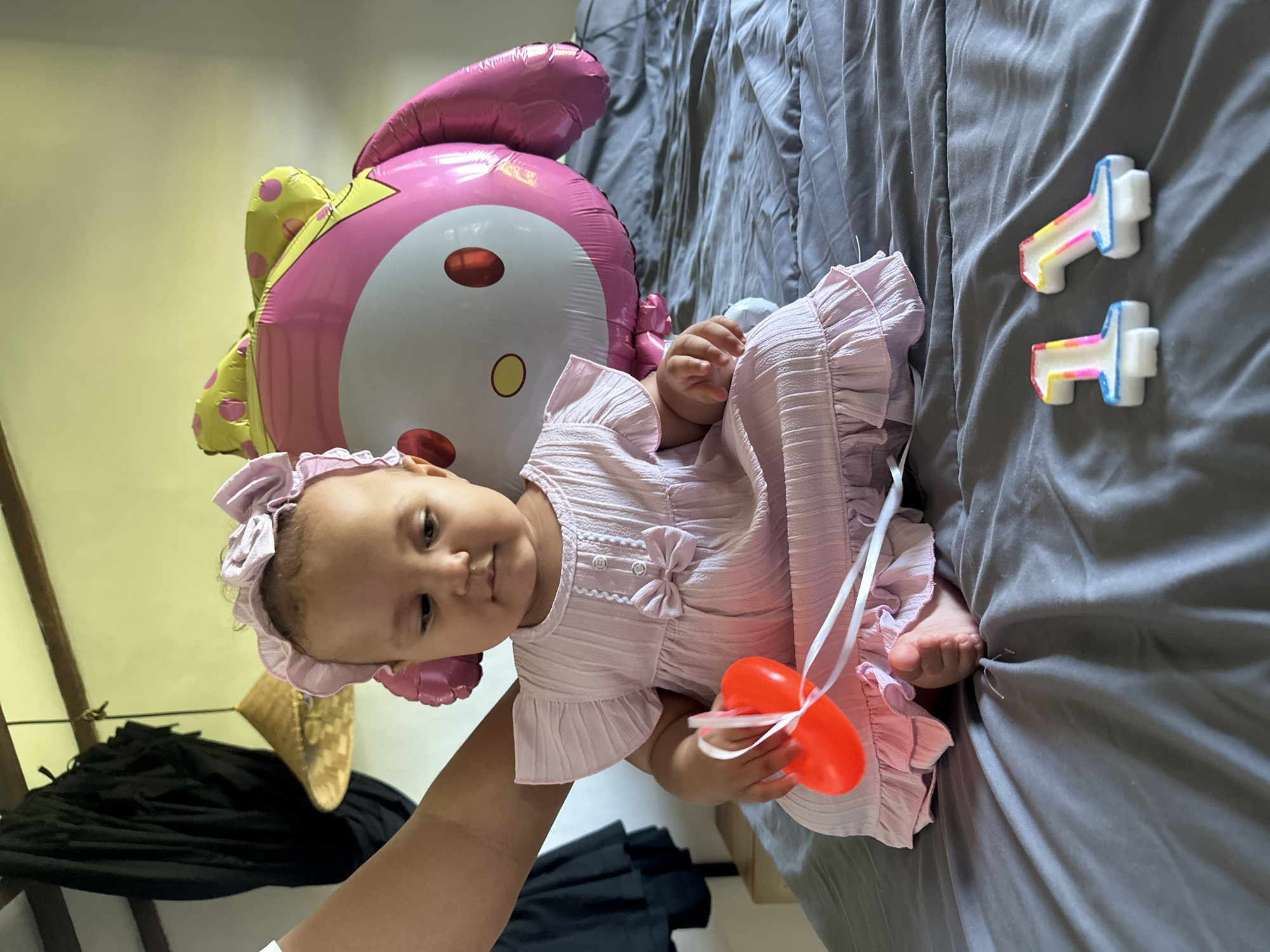 A baby girl in a pink ruffled dress and headband lies on a gray blanket. A large, pink balloon shaped like a cartoon character, possibly My Melody, is behind her. Her left hand holds a red, heart-shaped toy attached to a white ribbon. Two small, T-shaped candles with rainbow-colored edges are placed on the blanket to the right of the baby. The scene appears to be indoors, likely a bedroom, given the presence of the blanket and the soft lighting. The overall mood is celebratory, suggesting a birthday or special occasion for the child. There is no visible text in the image. The location is identified as North Kuta, Indonesia.