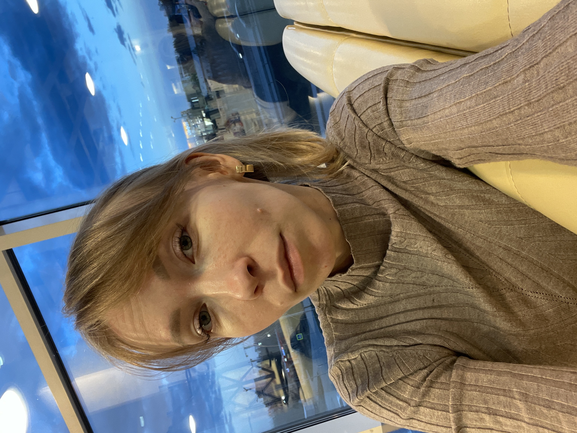 A woman with light skin and blonde hair is facing the camera. She is wearing a ribbed sweater and a gold rectangular earring is visible on her left ear. She appears to be seated on a light beige or yellow cushioned seat. Behind her, a window reflects an urban environment with a twilight sky. The reflection shows buildings, lights, and sky, suggesting it is dusk or dawn. The image appears to be taken indoors. Though it is difficult to place exactly, the architecture and light could support the conclusion that this photo was taken somewhere in the city of Perm, Russia.