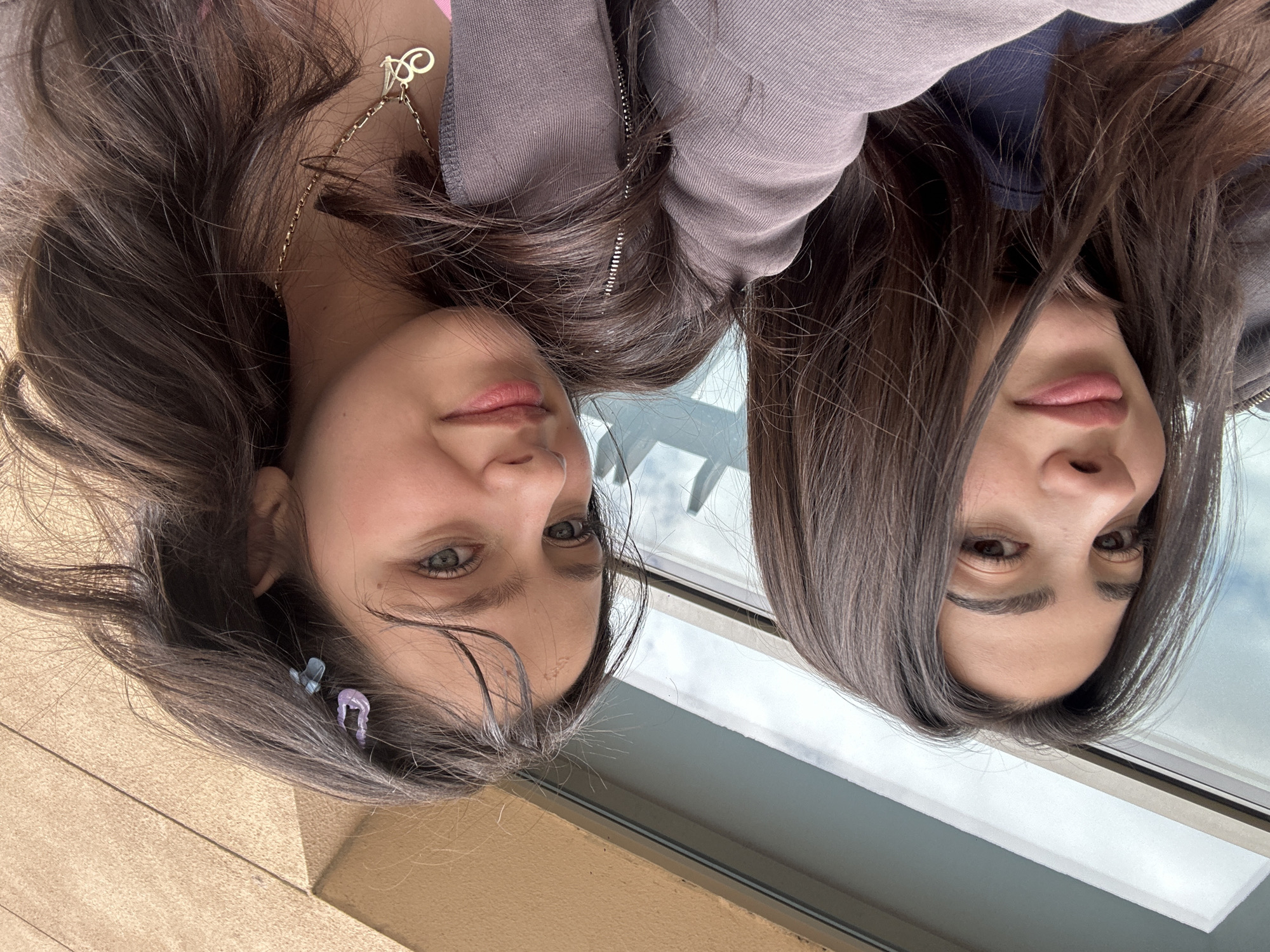 Two young women are positioned close together, facing the camera, with the image rotated 180 degrees. They appear to be indoors near a window, with a view of Yerevan visible through the glass. The woman on the left has light eyes and two decorative hair clips in her hair. She is wearing a gold necklace. The woman on the right has dark eyes and hair. Both have similar skin tones and lip colors. The environment suggests an interior setting in Yerevan, indicated by the window view.