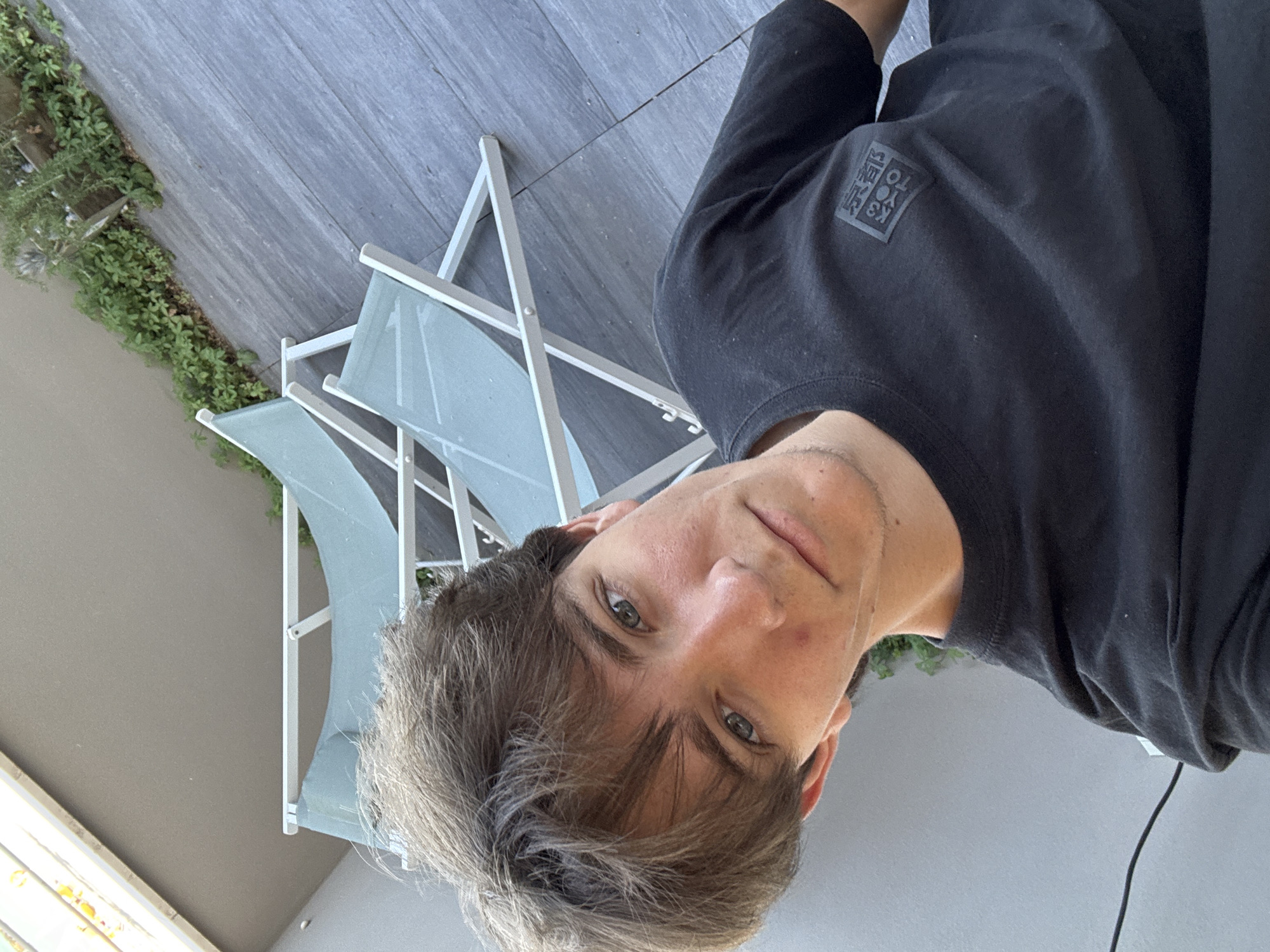 A young Caucasian male with light brown, tousled hair and green eyes is captured in a selfie taken outdoors. He is wearing a black t-shirt with a rectangular, dark-colored emblem on the left chest area. The emblem appears to contain Japanese characters within a box shape. The background features a paved deck, a lush green hedge, and two light blue lounge chairs with white frames. A light-colored wall is visible on the left side of the frame. The lighting suggests it is daytime, possibly outdoors on a balcony or patio. The location is identified as Cupra Marittima, Italy.
