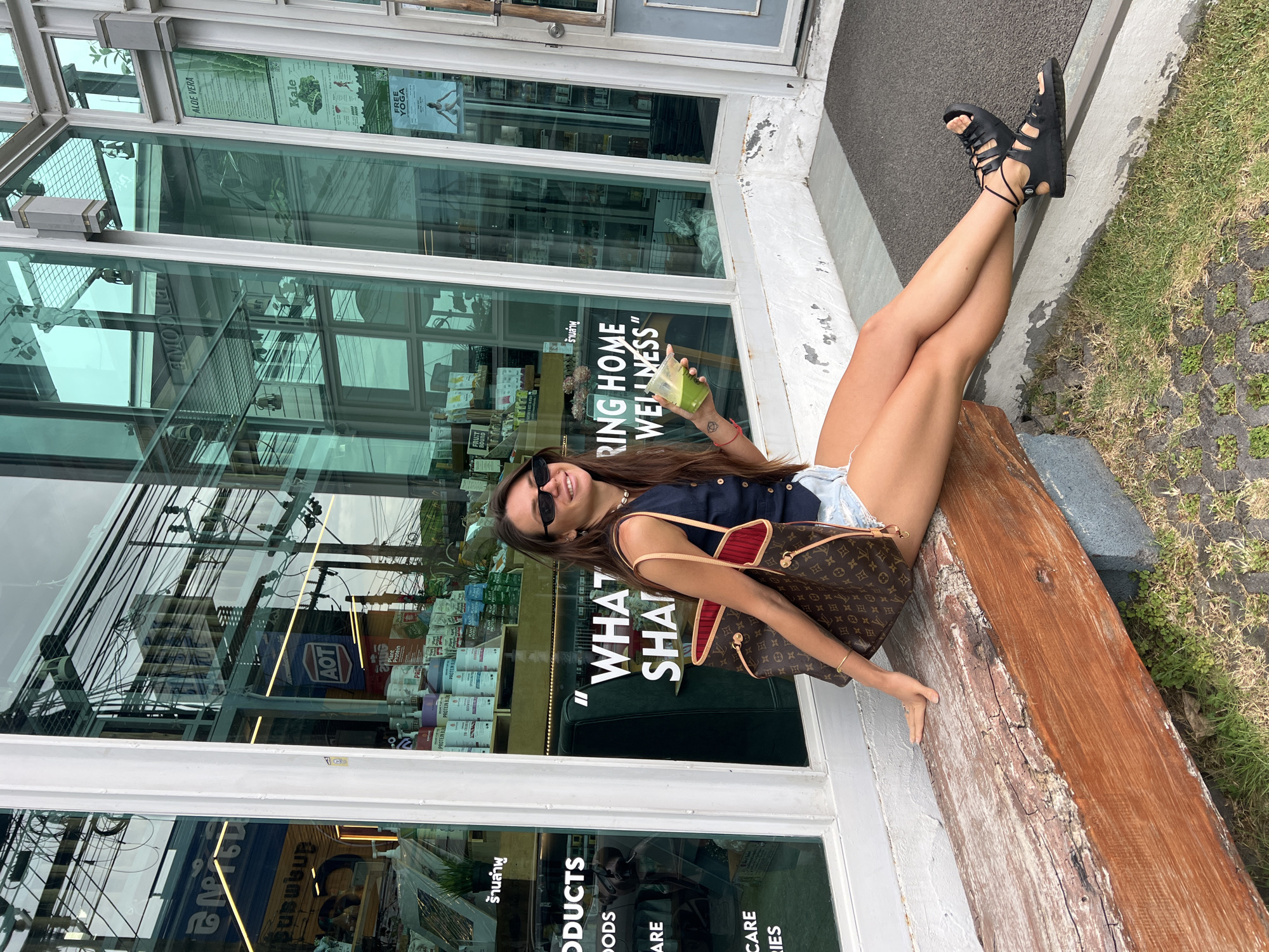 A young woman is captured in a full-body shot, smiling broadly at the camera while sitting on a rustic wooden bench outside a glass-fronted establishment in Baan Lamai, Thailand. She is holding a clear cup with a vibrant green drink. Dressed in a dark sleeveless top, light denim shorts, and black lace-up sandals, she appears relaxed, with sunglasses on her head. A large brown tote bag with a distinct pattern is placed next to her on the bench.
The building behind her features a modern design with expansive glass windows and white frames, reflecting the bright outdoor environment, including power lines and other distant structures. Through the windows, shelves stocked with health and wellness products, such as protein powders ("PROTEIN BE", "PROTEIN BL", "Plant Protein") and "FRUIT BOUND" items, are visible. Various signs on the glass convey the store's focus, including phrases like "RING HOME WELLNESS," "WHAT SHA," "PRODUCTS," "FOODS," "CARE," "LIES," "ALOE VERA," "Kale," and "FREE YOGA." Thai script "ร้านลำพู" (Raan Lamphu) is clearly visible, identifying the venue, along with a partial "日文版" (Japanese version) and an "AOT" logo reflected. The immediate surroundings include dark ground paving and sparse green grass, indicating an outdoor seating area. The sunny weather and the woman's cheerful expression contribute to a positive and inviting atmosphere.