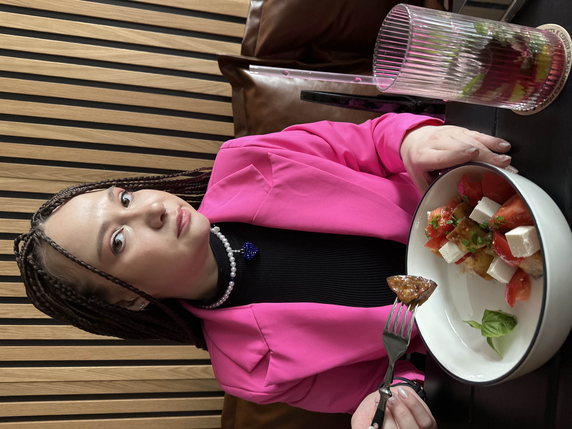 The image portrays a young adult female seated at a dark table indoors, located in Saratov, Russia. She looks directly at the viewer with a neutral expression. Her hair is styled in numerous small, dark braids. She wears a bright pink blazer over a black ribbed top and a pearl necklace featuring a central blue beaded heart pendant. A small nose piercing is visible. Her left hand holds a metal fork with a piece of dark-brown, likely fried, food item. This food item is positioned above a white bowl with a black rim, containing a salad composed of diced red tomatoes, white cuboid feta cheese, and green basil leaves. To the right of the bowl, a tall, clear, vertically ribbed glass holds a reddish beverage with green mint leaves and yellow citrus wedges, resting on a coaster. The background consists of a wall featuring alternating vertical light wooden panels and dark strips, with a segment of a brown leather-padded seat visible on the right.