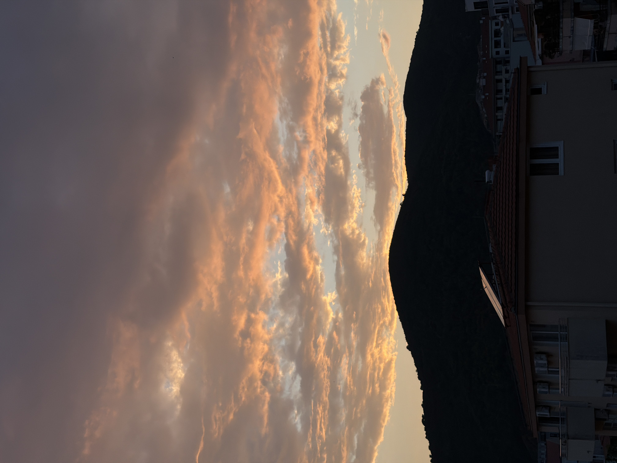 An underexposed, vertical shot captures a silhouetted mountain range against a dramatic sunset sky over Budva, Montenegro. The sky is filled with billowing clouds, painted in hues of peach, orange, and gray, with slivers of pale blue peeking through. The dark, undulating line of the mountains occupies the lower third of the frame. In the foreground, the dark silhouettes of buildings are visible, with tiled roofs and windows. One building in the foreground has solar panels on its roof. The overall impression is one of a serene, yet dramatic, end to the day.