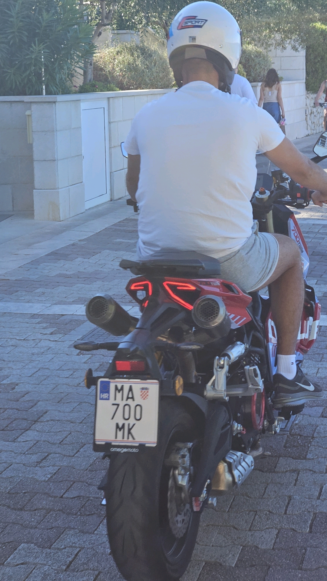 A man wearing a white t-shirt, grey shorts, white socks, and black athletic shoes is riding a red and black motorcycle. He is wearing a white helmet with a red and blue stripe and the word "SPORT" on it. The motorcycle has a white license plate with "HR MA 700 MK" written on it, along with a small Croatian flag. The motorcycle also has dual exhaust pipes and red taillights. The man is on a paved road with a brick pattern. In the background, there is a stone wall, a white door, and green bushes. The scene appears to be in a city or town, likely Općina Tučepi, Croatia, given the license plate.