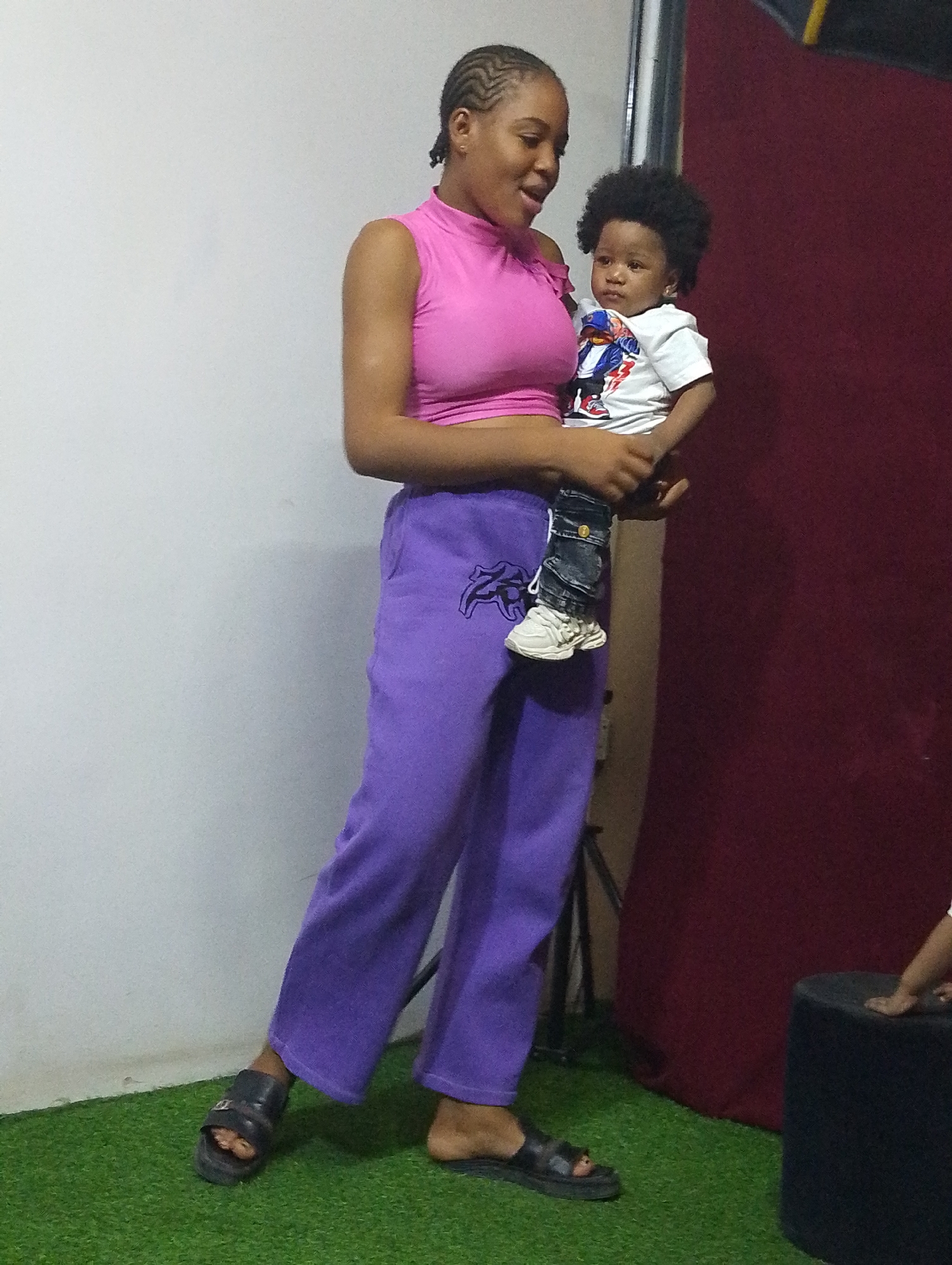 A young Black woman is pictured holding a baby indoors, likely in a makeshift photo studio or designated photo area. The woman, with braided hair, is wearing a pink sleeveless turtleneck crop top, loose purple sweatpants with a dark logo on the left thigh, and black slide sandals. She is looking down at the baby with a gentle, open-mouthed expression, suggesting she is speaking or smiling.
The baby, appearing to be an infant, has dark, curly hair and is dressed in a white short-sleeved t-shirt with a colorful graphic print (possibly a cartoon character), denim shorts or jeans, and white sneakers. The baby is looking slightly to the left with a calm, neutral expression.
The background features a plain white wall on the left and a dark red fabric backdrop on the right. The floor is covered with green artificial turf, giving the impression of an outdoor setting indoors. A black stool or low prop is visible on the right, and a black metallic stand (possibly a light stand or tripod) can be seen behind the woman's legs. The scene is well-lit, suggesting a planned photo session. The interaction between the woman and baby conveys a tender and affectionate moment. The location context is Babban Giji, Nigeria.