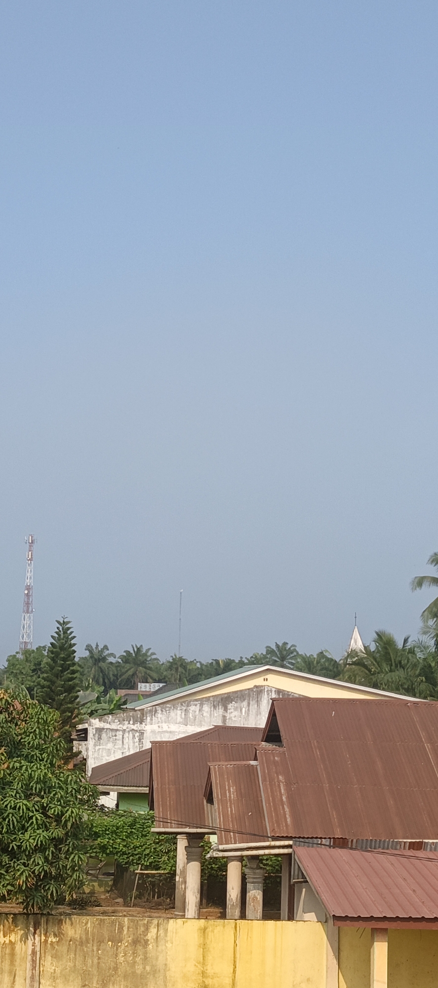 The image captures a view of buildings and greenery under a clear blue sky, likely in Negeri Lama, Indonesia. In the background, a tall, red and white telecommunications tower stands prominently amongst lush trees. Further to the right, a smaller, slender tower is visible, along with the pointed spire of what appears to be a church, also surrounded by foliage. The foreground is dominated by the roofs of several buildings. The most prominent are a row of rusted, corrugated metal roofs with triangular peaks, supported by classical-style pillars. Behind these, a building with a light yellow painted exterior and a white, textured wall is visible. The lower portion of the image shows a thick, yellow-painted concrete wall or structure, providing a base to the scene. The overall impression is of a semi-urban or semi-rural environment with a mix of functional structures and natural elements.