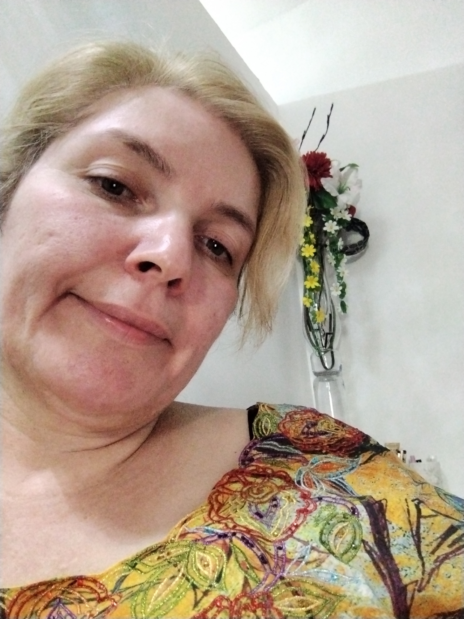 A blonde-haired woman with fair skin is pictured from a low angle, looking directly at the camera with a slight smile. She is wearing a colorful, patterned garment that covers her shoulder. Behind her, affixed to a white wall, is a glass vase holding an arrangement of artificial flowers in red, white, and yellow. The location is not discernible, and there is no information in the image to establish that this image was taken in Al Mas'oudi, United Arab Emirates.