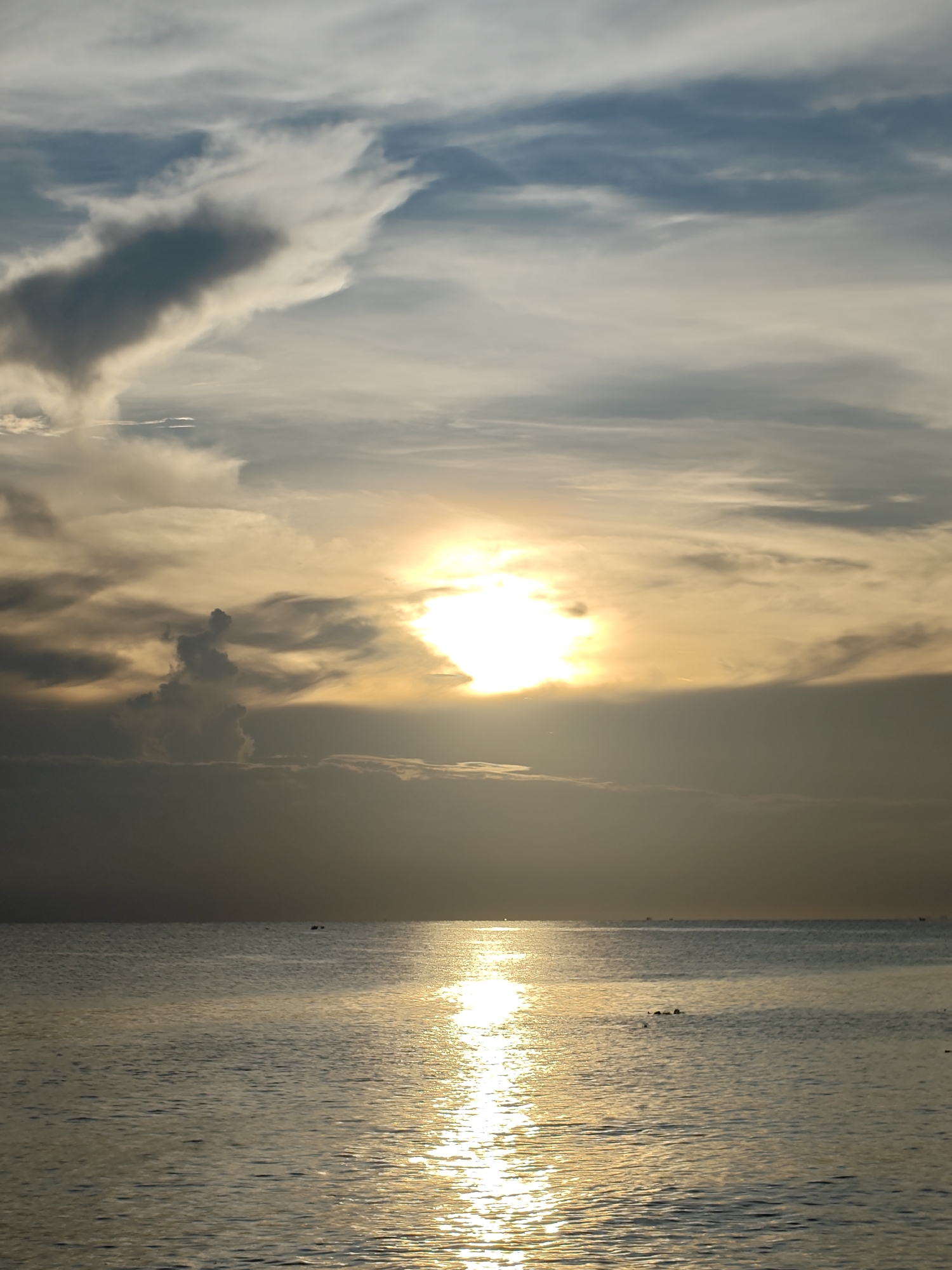 The image depicts a sunset over the sea in Lhokseumawe, Indonesia. The sun is partially obscured by clouds, casting a golden glow over the water. The sky is a mix of blue, gray, and white clouds. The sea is relatively calm with a bright reflection of the sun stretching towards the viewer.