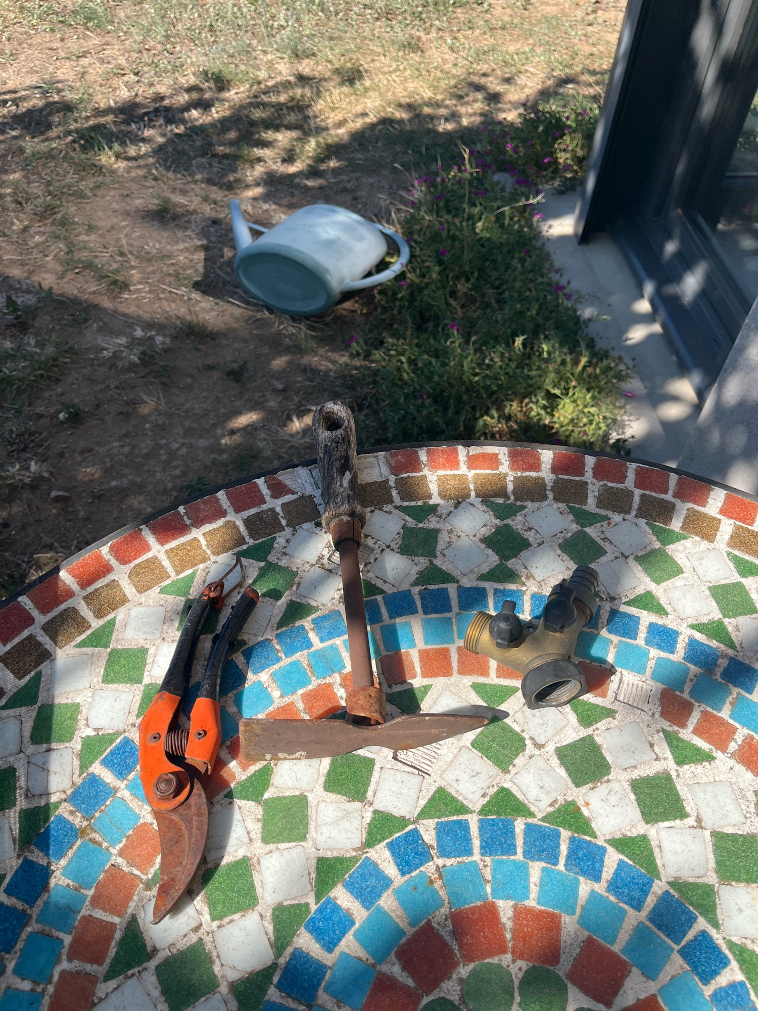 In Chambœuf, France, an outdoor scene features a mosaic-tiled table. On the table are gardening tools including orange-handled pruning shears and a trowel with a wooden handle. A brass Y-shaped hose splitter with two black valves is also present. In the background, a light blue plastic watering can lies on its side on dry, grassy ground, adjacent to a patch of small purple flowers and a dark grey structure that appears to be a building or fence. The lighting suggests it is daytime, with shadows cast by unseen objects.