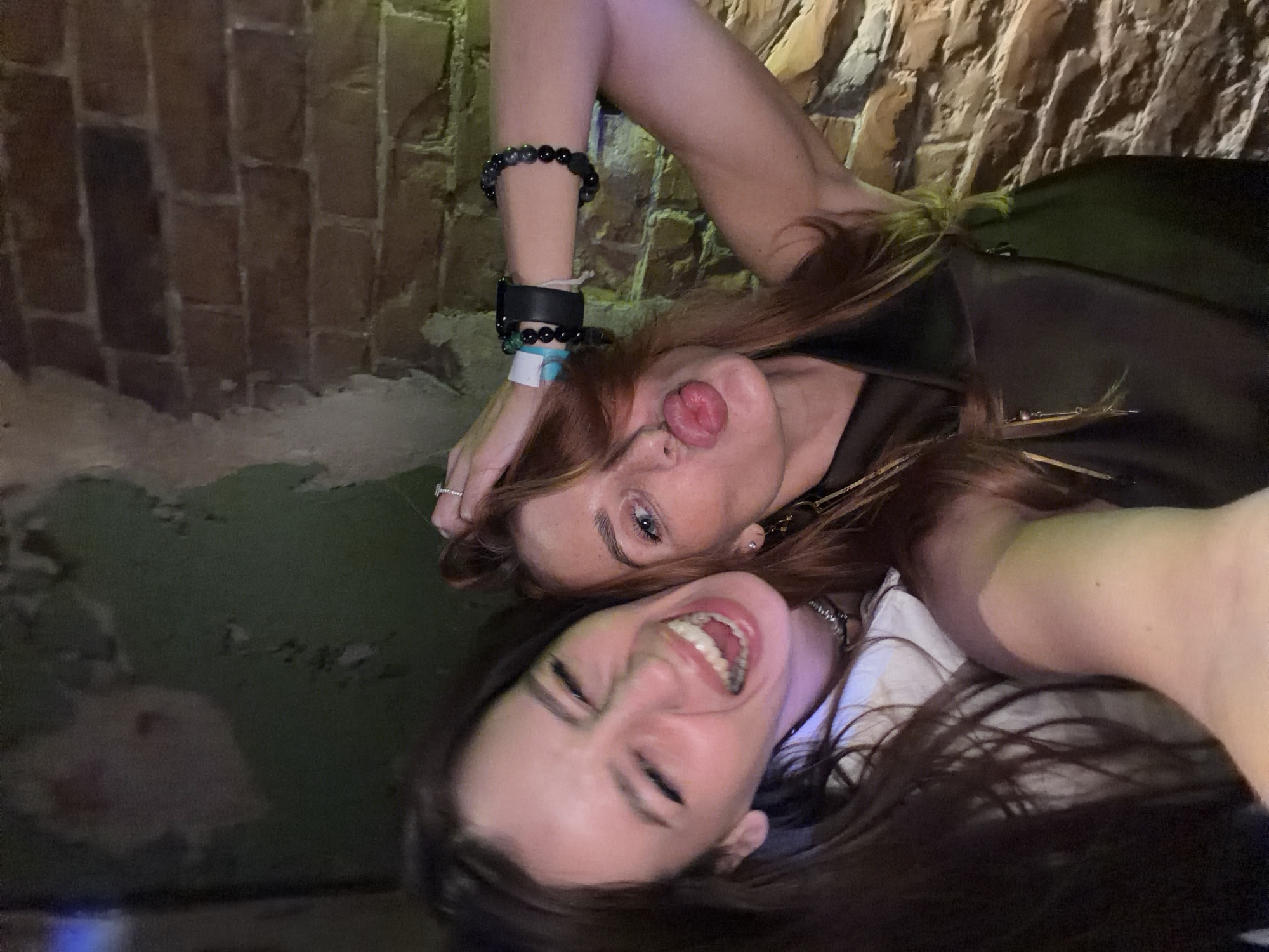 Two women are lying down next to each other, facing the camera. The woman on top has red hair and is sticking her tongue out. She is wearing multiple bracelets and a black smart watch on her left wrist. The woman on the bottom is smiling broadly, showing her teeth, and has dark hair. The background consists of a brick wall and a dark, indistinct surface. The location is in the city of Unknown, Unknown.