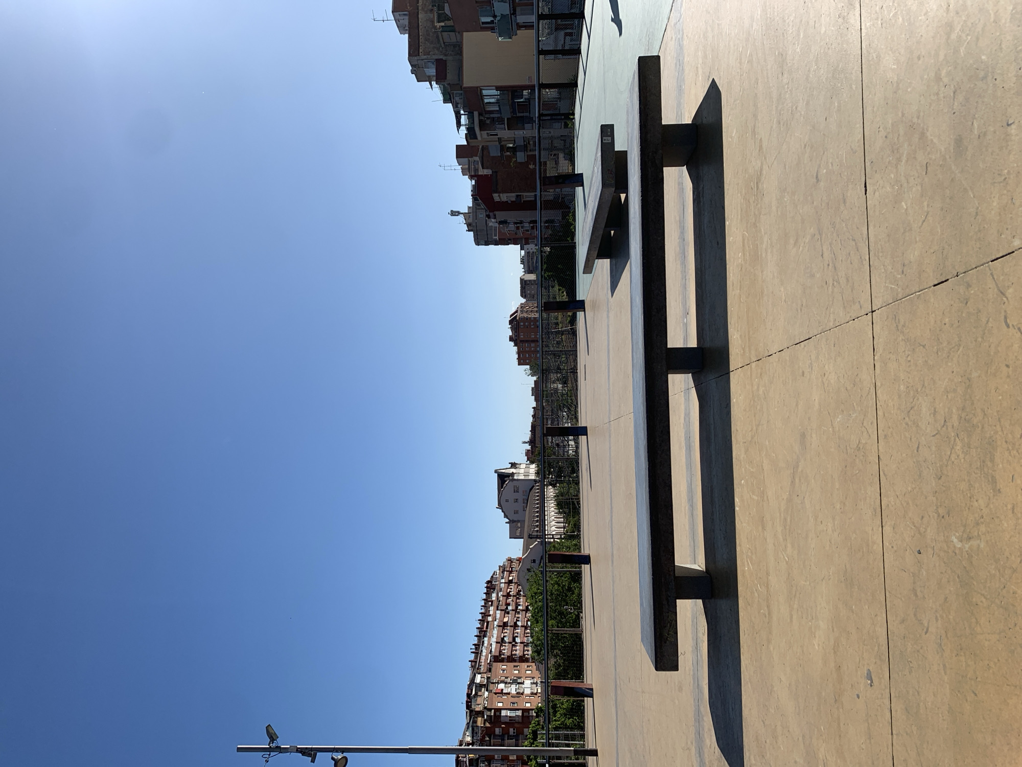 The image depicts a cityscape in l'Hospitalet de Llobregat, Spain. The view is taken from a high vantage point, revealing a row of buildings against a clear blue sky. A concrete platform occupies the foreground, featuring a dark-colored bench with metal supports. A streetlight fixture is visible in the lower-left corner. The buildings exhibit varied architectural styles and colors, suggesting a mixed residential area.