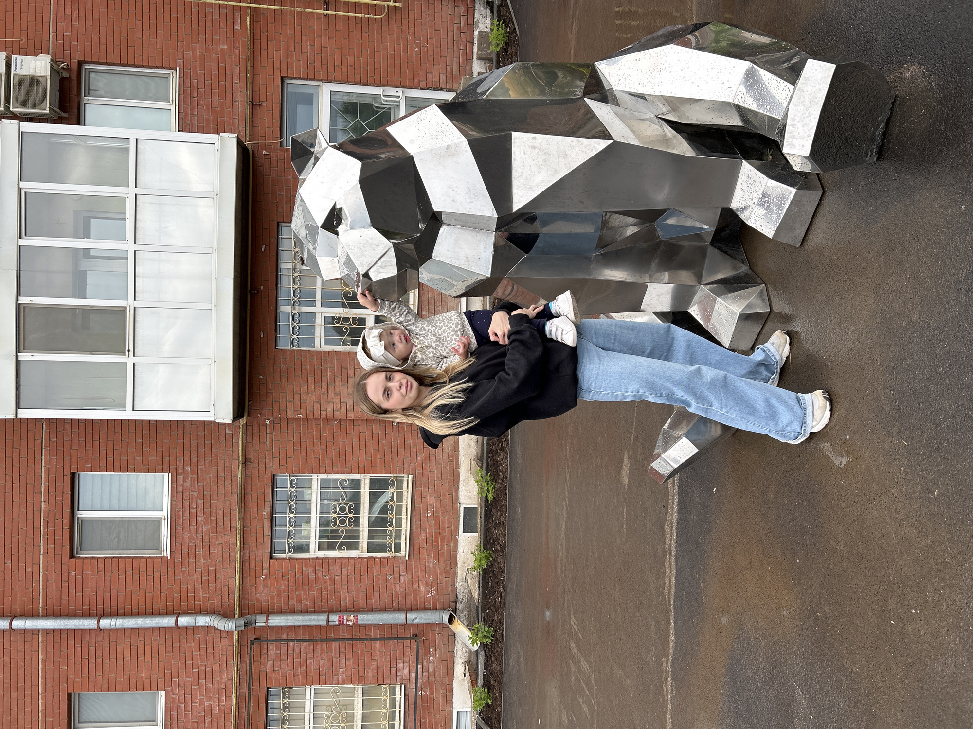 A young woman is depicted holding a small child next to a large, reflective, geometric metallic sculpture, likely in an urban or residential setting in Азинский, Russia. The woman, with long blonde hair, wears a black hoodie, light blue jeans, and white sneakers, and looks towards the camera with a slight smile. The child, with blonde hair and a white headband, is dressed in a patterned top and dark bottoms, and is looking away from the camera.
The prominent sculpture is composed of numerous faceted, silver-toned metallic panels, giving it a polygonal, modern art appearance, possibly depicting a large animal like a bear. Its surface is dotted with water droplets, suggesting recent rainfall. The ground beneath is dark, wet pavement or asphalt.
In the background, a multi-story building with reddish-brown brick walls is visible. It features several windows; some have plain white frames, while others are adorned with decorative metal grates. A large, white or silver enclosed balcony structure with multiple glass panes is attached to the brick facade. Air conditioning units are mounted on the brick wall. Metal pipes run along the building's exterior. A narrow garden bed with sparse green plants separates the building from the paved area.
The scene captures a casual daytime moment outdoors, under overcast skies or after rain. No visible text is discernible in the image.