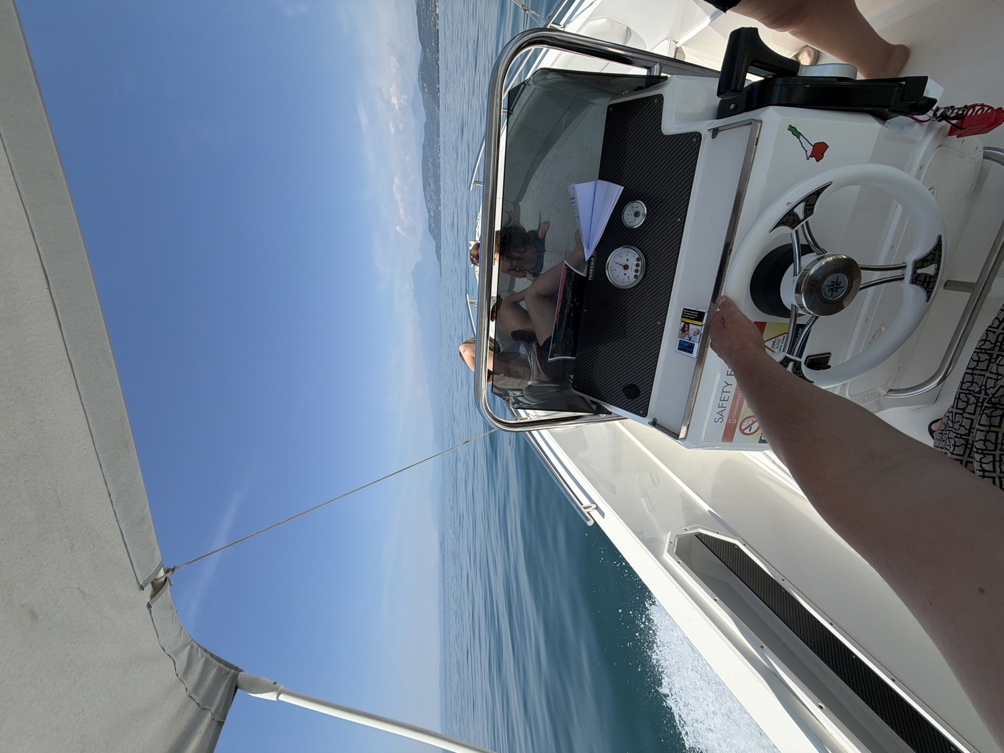 The image shows a point-of-view perspective from the helm of a white motorboat navigating on a calm body of water, likely Lake Garda, with the shoreline of Lazise, Italy, visible in the distance. The sky is clear blue. In the foreground, a person's arm and hand are gripping the white steering wheel. The boat's dashboard is visible, featuring gauges and a "SAFETY FIRST" sign. Behind the dashboard, two individuals are seated on the boat, one with their back to the camera and another facing away. The boat is moving, creating a white wake behind it. The overall scene depicts a recreational boat trip on a sunny day.
