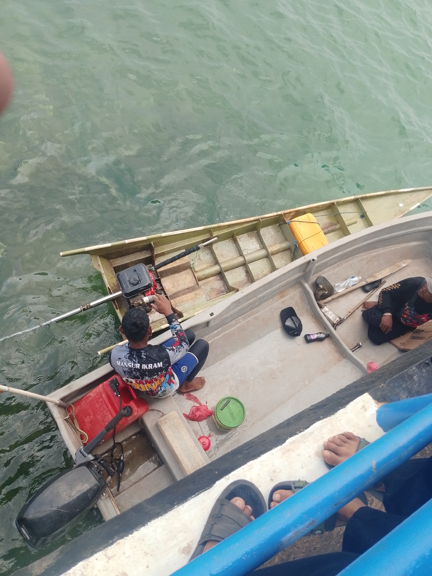 In Batam, Indonesia, two men are in a small aluminum boat docked alongside a larger vessel. One man, wearing a black and colorful long-sleeved shirt and blue shorts, sits in the front of the boat, appearing to be adjusting a black outboard motor. He has dark hair and is barefoot. Next to him is a red fuel can. In the rear of the boat, another man, wearing a black shirt and a cap, is seated. Various items are scattered in the boat, including a yellow container, a paddle, a water bottle, and footwear. The boat is on the water, which appears slightly murky and green. The context suggests a potential fishing or transport scenario.