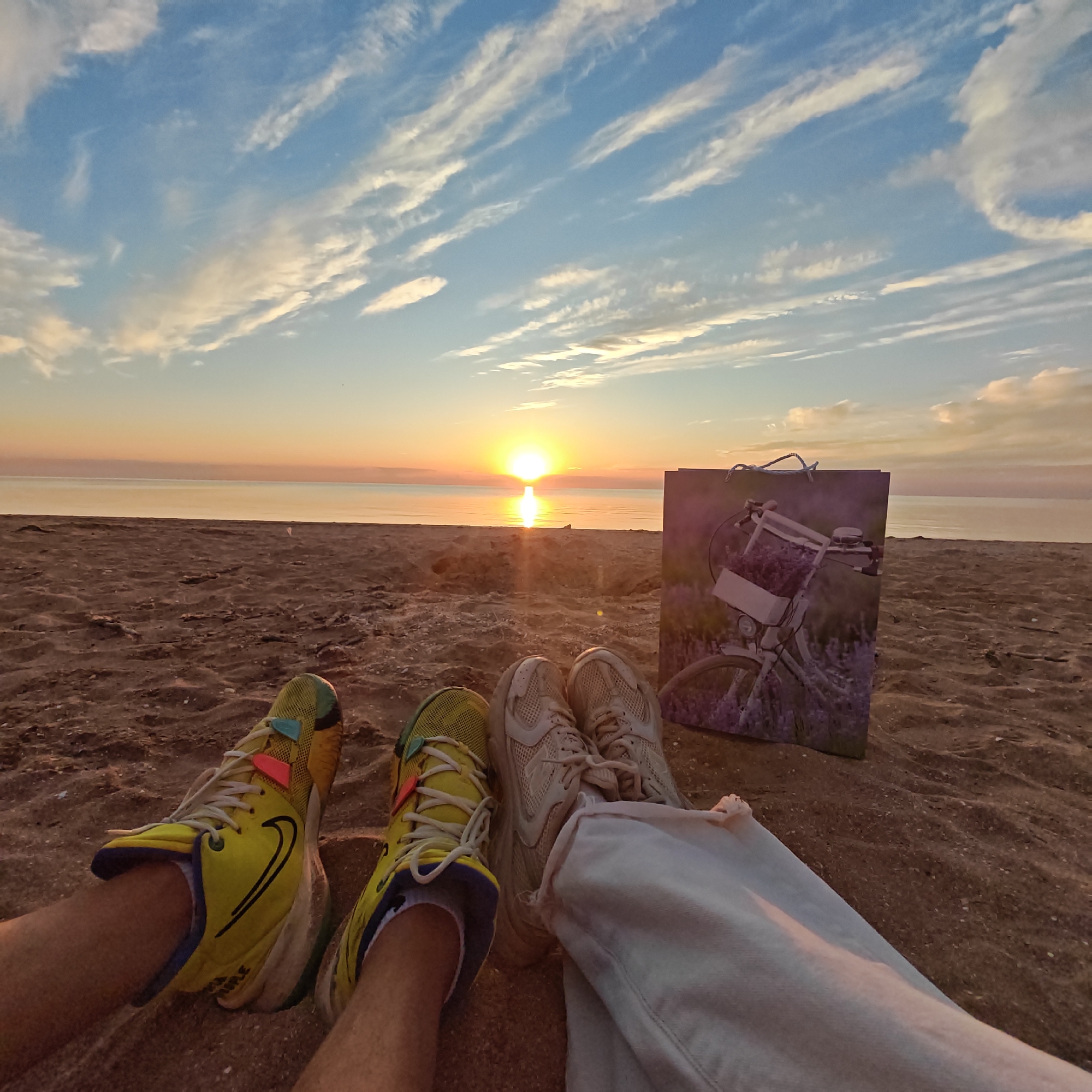 The image depicts a low-angle view of two individuals' lower legs and feet on a sandy beach, facing the sea during a sunset or sunrise in Unknown, Unknown.
The horizon is dominated by a bright, round, orange-yellow sun reflecting a strong column of light on the calm, still water. The sky above is a gradient of warm orange and yellow near the horizon, transitioning to light blue, populated by wispy, elongated white clouds extending across the frame.
The foreground consists of dark brown sand, textured with small ridges and sparse debris. To the left, a person's left leg and foot are visible, wearing a bright yellow athletic shoe with a black Nike swoosh logo, multi-colored accents (blue, pink, green), and white laces. The shoe has a thick, light-colored sole. Adjacent to this, to the right, are the feet of a second individual. This person wears light-colored, possibly off-white, athletic shoes with textured mesh uppers and thick soles. Their lower legs are covered by light-colored, presumably white or off-white, trousers or jeans, which appear slightly distressed near the knee.
To the right of the individuals' feet, a purple shopping bag stands upright on the sand. The bag features a printed image of a white bicycle with a front basket filled with lavender flowers. Blue braided rope handles are visible at the top of the bag. The overall scene captures a tranquil moment at the beach, observing the sun setting or rising over the water.