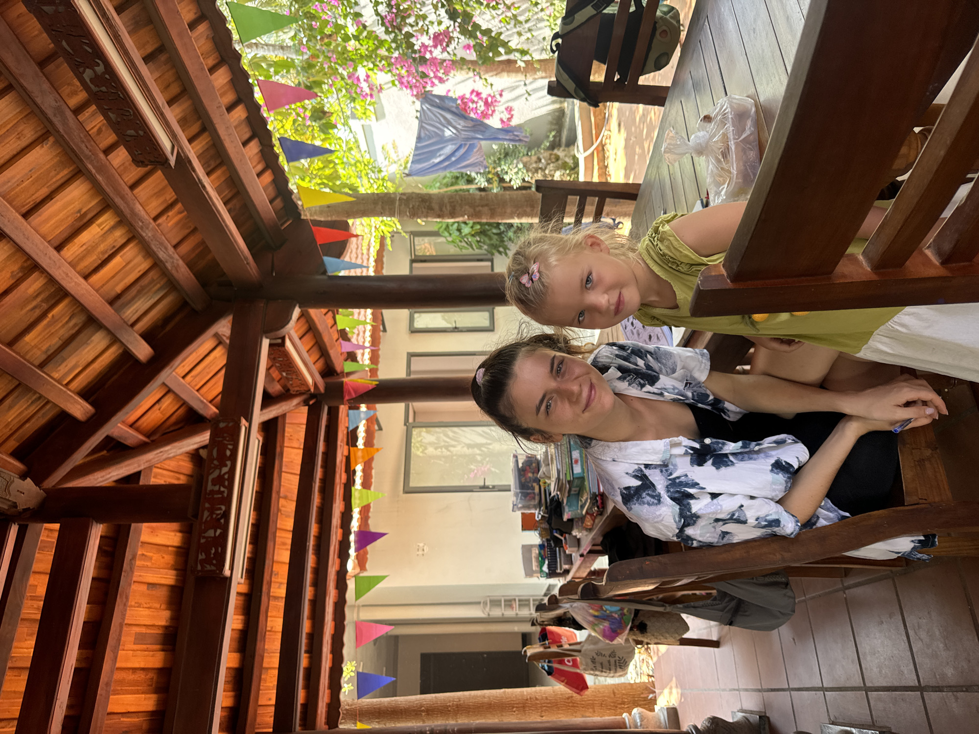 A woman and a young girl are seated at a wooden table under an elaborate wooden roof structure decorated with colorful pennant banners, possibly in Mũi Né, Vietnam. The girl, with blonde hair, wears a yellow dress. The woman is in a patterned shirt. The setting appears to be an open-air cafe or restaurant. A backpack sits on a chair in the background. A bag of food rests on the table. The environment includes lush vegetation and other buildings visible beyond the roof. The image shows both the interior structure and the outdoor environment.