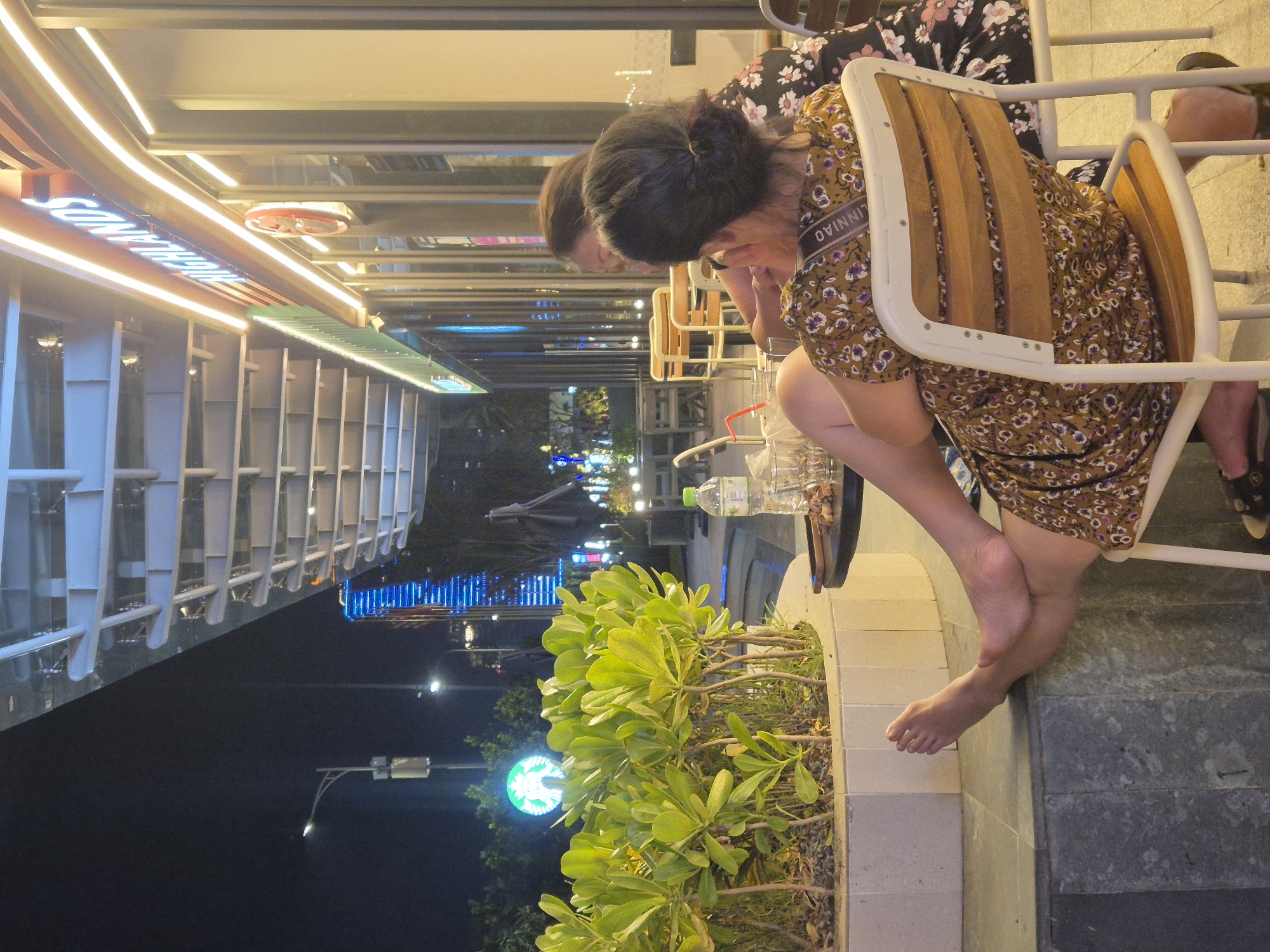 This media file shows two individuals sitting at an outdoor cafe or restaurant in Da Nang, Vietnam at night. The person in the foreground, a woman with dark hair pulled back, is wearing a floral dress and is seated on a white chair with wooden slats. Her legs and bare feet are visible as she sits with one leg bent and the other extended. She appears to be looking down, possibly at something on the table or her phone. Another person, partially visible in the background, is wearing a dark floral patterned top.
The setting is illuminated by ambient lighting from the establishment and streetlights. To the left, a building facade is visible with a curved structure illuminated by warm, white LED lights. Text is visible on this facade, reading "SONGCHINH". Further to the left, there are several levels of white structures that appear to be part of the building's exterior or interior. In the mid-ground, a tall building with blue lights is visible, suggesting an urban environment. In the foreground and mid-ground right, there is a planter box filled with lush green foliage. A bright green circular logo, resembling the Starbucks logo, is visible in the background, further indicating a commercial area. The overall atmosphere appears relaxed and casual, consistent with an evening outing at a cafe.