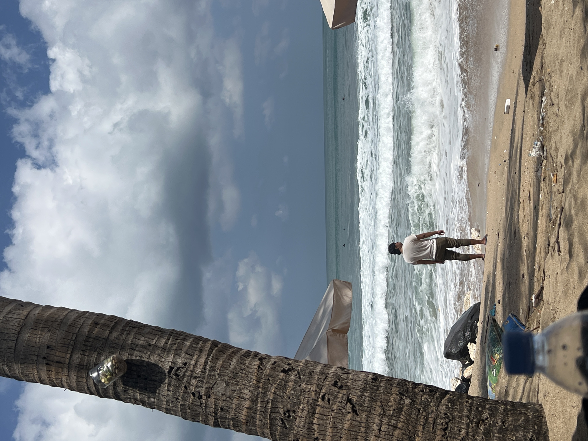 A man stands on a sandy beach in Kuta, Indonesia, facing the ocean. He is wearing a light-colored t-shirt and dark pants, with a black baseball cap. The ocean has moderate waves, with white foam breaking on the shore. In the foreground, a large palm tree trunk is on the left side of the frame. There are several beach umbrellas visible in the mid-ground. The sky is partly cloudy, with a mix of blue sky and darker clouds. There is some debris on the sand, including plastic bags and a partially visible water bottle in the lower left corner. The overall scene is one of a typical beach day, with a solitary figure enjoying the view of the sea.