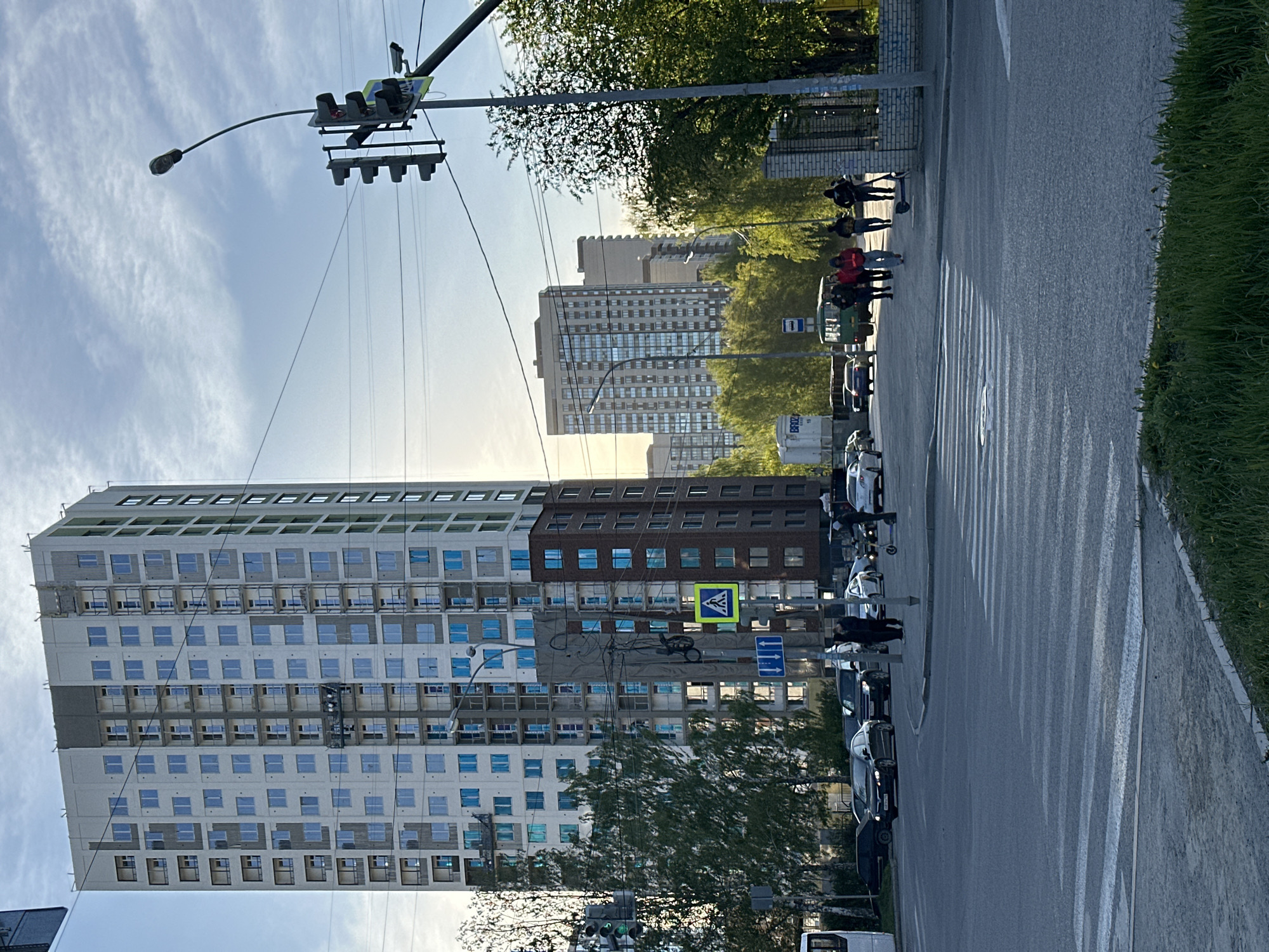 The image depicts a street scene in Yekaterinburg, Russia. A tall building is under construction. Pedestrians are crossing the street at a crosswalk. Cars are parked along the side of the street. A traffic light is visible. The weather appears to be sunny.