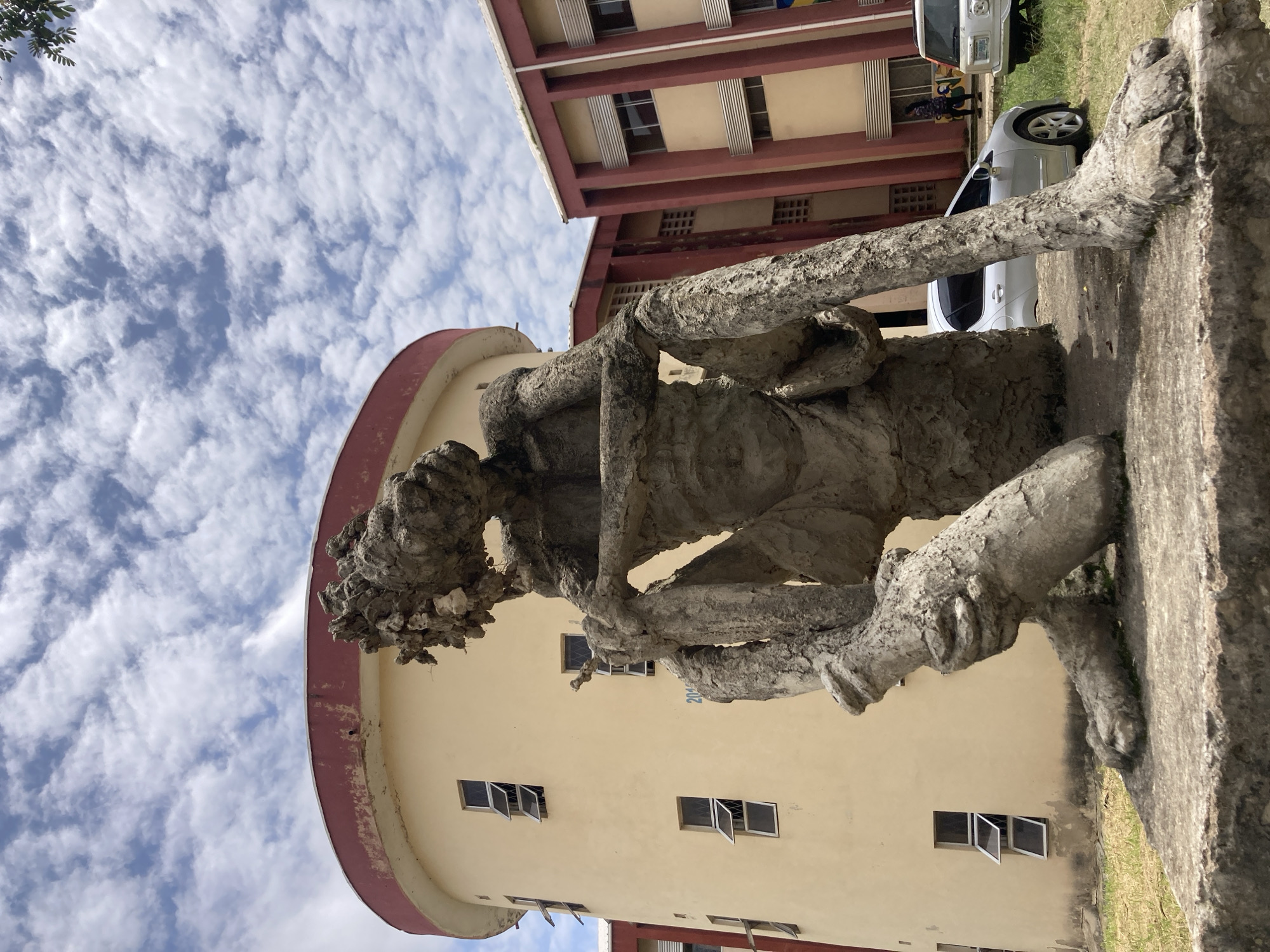 A low-angle, full outdoor shot shows a large, rough, textured concrete sculpture of a seated, contemplative figure. The sculpture is a muted gray and appears to be made of rough-hewn cement. The figure is hunched over, with its head bowed and its arms resting on its knees, its face obscured. It is positioned on a concrete pedestal in front of a pale yellow, cylindrical building with a red trim and several open windows. To the right of the cylindrical building is a red and cream-colored rectangular building with multiple windows. In the background, there are two cars, a silver sedan and a white SUV, parked on a grassy area. The sky above is a bright blue with scattered white clouds. The image appears to be taken during the daytime. The context provided suggests this scene is located in the city of Rafin Par, Nigeria.