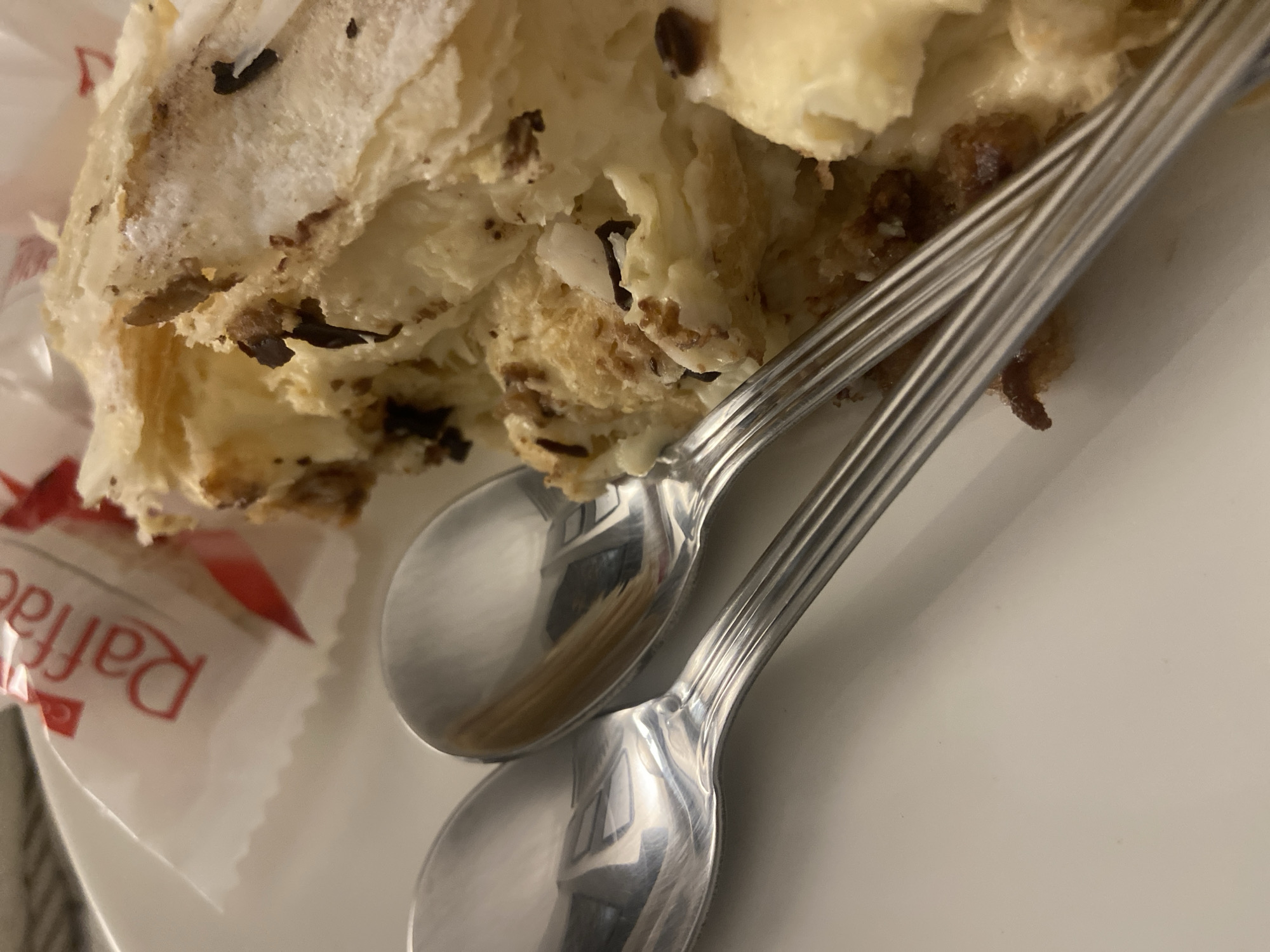 An indoor scene in Budva, Montenegro, depicts a partial view of a white ceramic plate holding a dessert, two spoons, and a candy. The dessert, situated on the right, is a layered pastry with visible flaky dough, a white or cream-colored filling, and numerous dark brown or black fragmented inclusions. It appears partially cut or broken. To the left of the dessert, two silver-colored spoons with ridged handles lie parallel on the plate. In the upper left background, a portion of a white candy wrapper with red text, identifying "Raffaello," is visible. Reflections on the spoons suggest an indistinct interior environment, possibly showing a window or light source. No specific landmarks or architectural features are present to pinpoint an exact location within Budva.