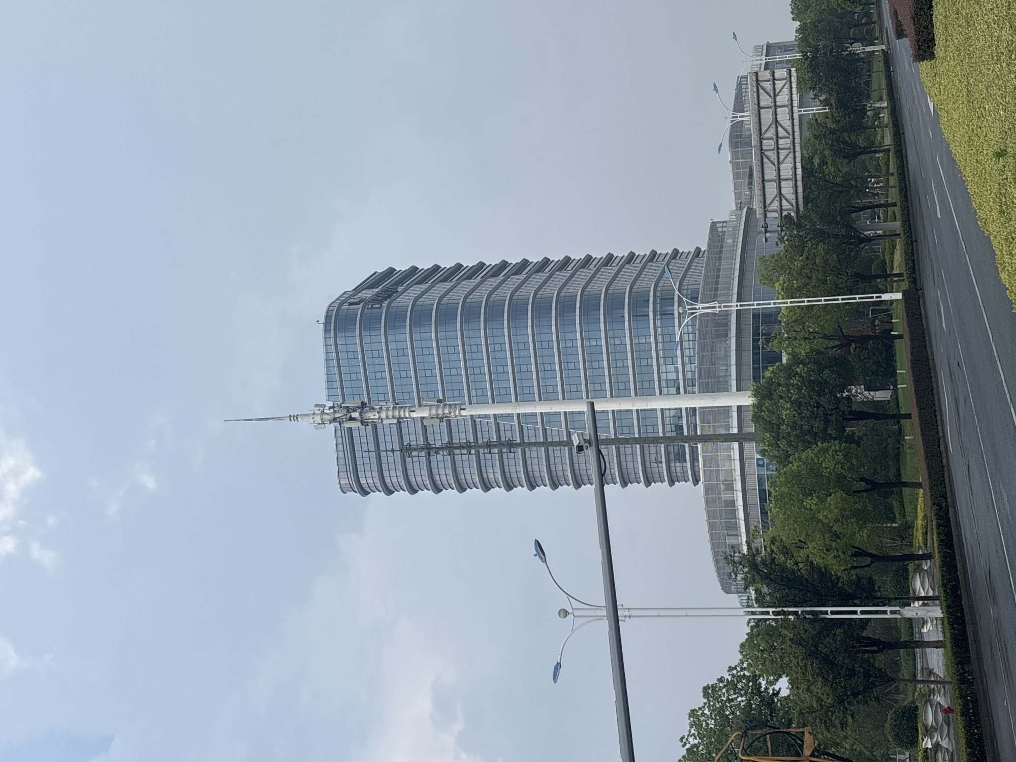 The image captures a view of a modern, curved glass office building in Suzhou Industrial Park, China, under a cloudy sky. A tall communications tower with antenna equipment is prominently positioned in front of the building. A street with wet asphalt and road markings runs in the foreground, bordered by a median with green bushes and trees. Several streetlights and a traffic light structure are visible along the roadside. A pedestrian bridge or elevated walkway connects to a lower circular section of the office building complex. Lush green trees and manicured landscaping surround the building and along the street. There are no visible people in the frame.