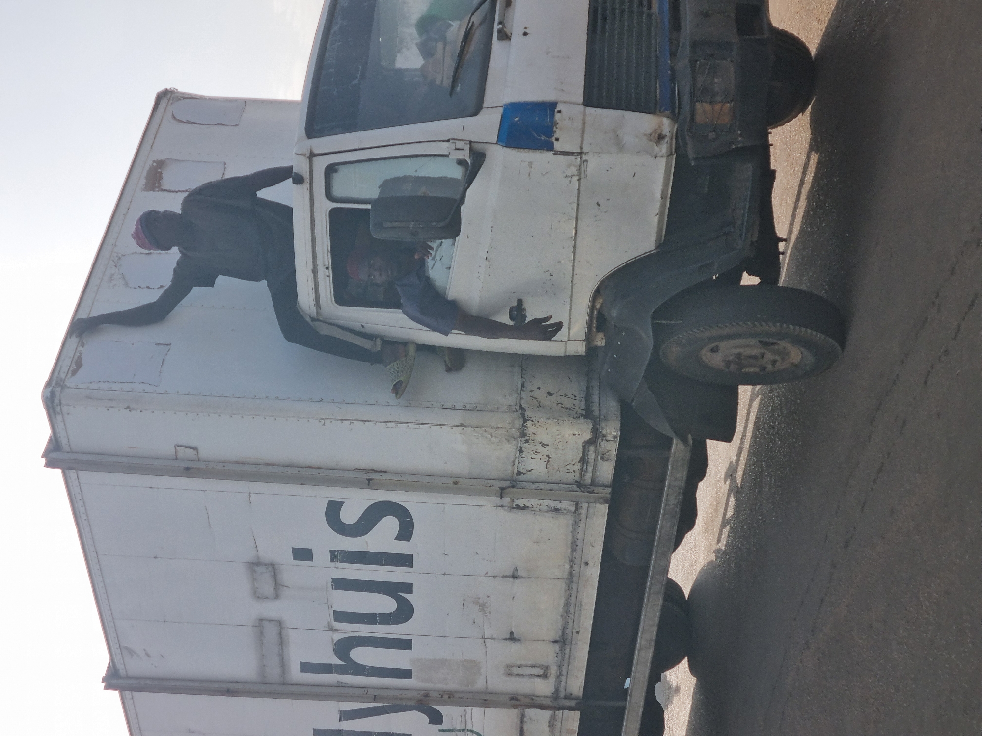 A white "lyhuis" branded truck is depicted on a paved road in Jos, Nigeria. One man is hanging off the side of the truck while another man is seated in the driver's seat. Both men appear to be of African descent.