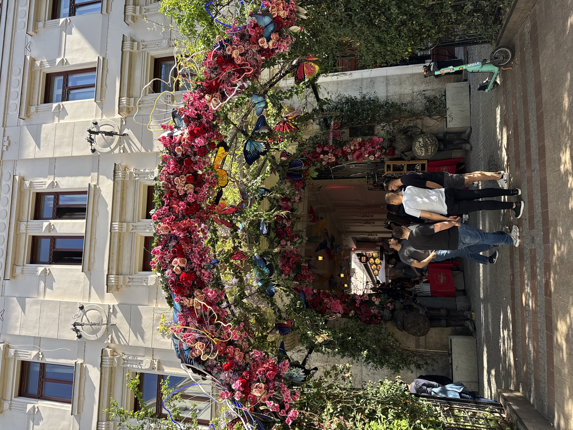 An outdoor street scene in Budapest, Hungary, captures a sunny day with a prominent, richly decorated archway spanning across the view.
On the left, a multi-story, cream-colored building with classic architectural details features rectangular brown-framed windows and decorative elements on its facade. A black, ornate metal fixture, possibly an old sign holder or clock, is mounted on the building between two windows.
The central feature is an elaborate archway, densely adorned with a vibrant display of artificial red and pink flowers, interspersed with green foliage. Numerous colorful artificial butterflies in shades of blue, yellow, pink, and orange are attached to the arch, along with some integrated neon light elements. This decorative structure appears to mark an entrance or a special area.
On the right, a paved sidewalk is occupied by several pedestrians. In the foreground, two men are visible from behind; one wears a white t-shirt and black pants, the other a black t-shirt and blue jeans. Further down the sidewalk, a light blue "BOLT" brand electric scooter, with "BOLT" and "COOL" visible on its side, is parked.
Underneath the floral archway, behind the people, are various commercial displays. A circular sign clearly reads "GOSPODA U WŁADKA" and "KUCHNIA POLSKA," indicating a Polish restaurant. Dark statues or figures and market stalls with red coverings are also present. The bright sunlight casts distinct shadows, suggesting it is either morning or afternoon on a clear day.