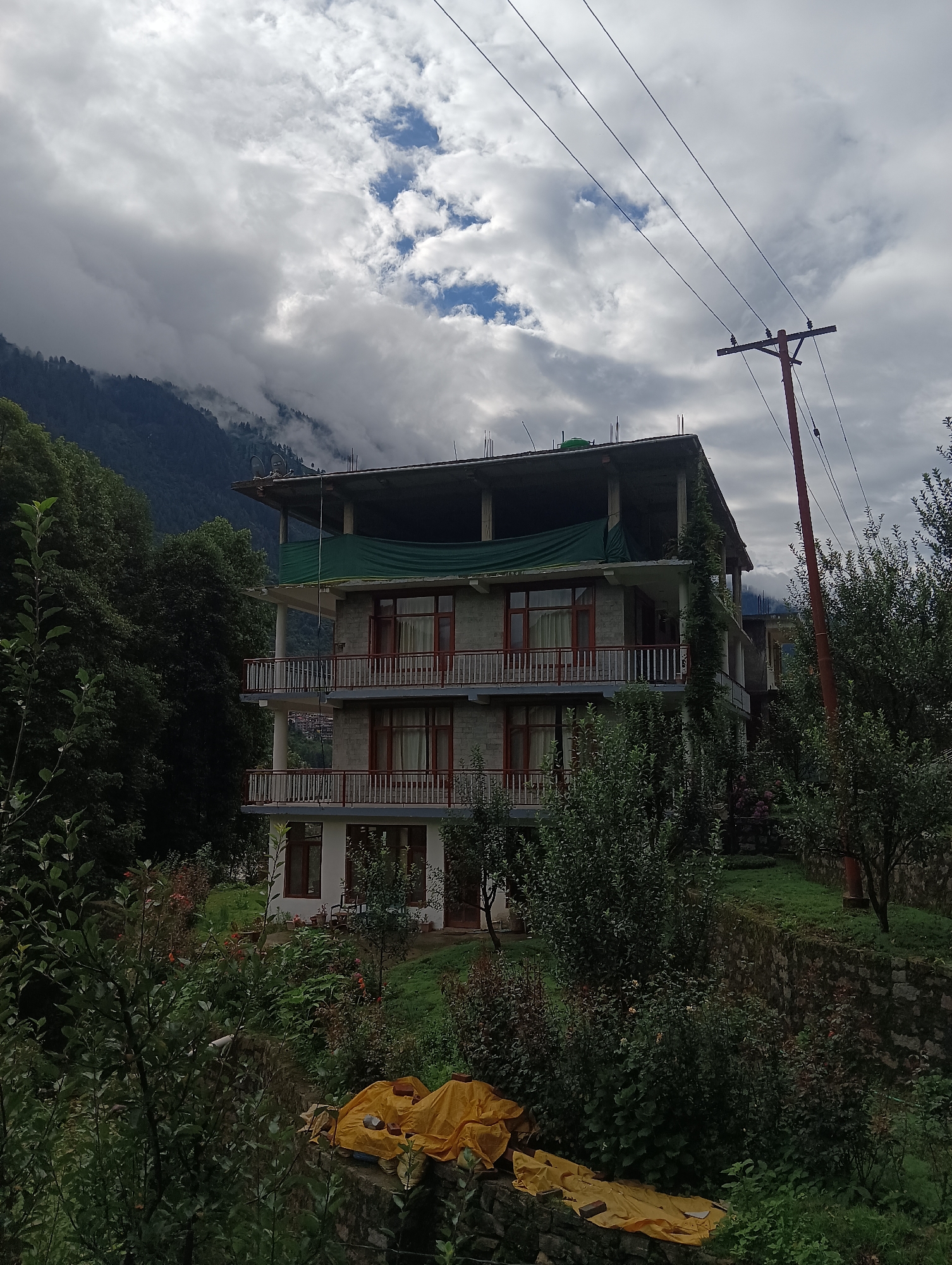 The image shows a multi-story building in Goshal, India, nestled amidst lush greenery and a mountainous backdrop. The building, likely a hotel or guesthouse, features balconies on each level with railing details. The architecture suggests it is made of stone with wooden window frames. The sky above is overcast with dramatic clouds. In the foreground, dense foliage and trees surround the structure, with a stone wall visible at the bottom of the frame. A power line pole with wires stretches across the upper right portion of the image. The overall scene conveys a sense of a rural, natural setting.
